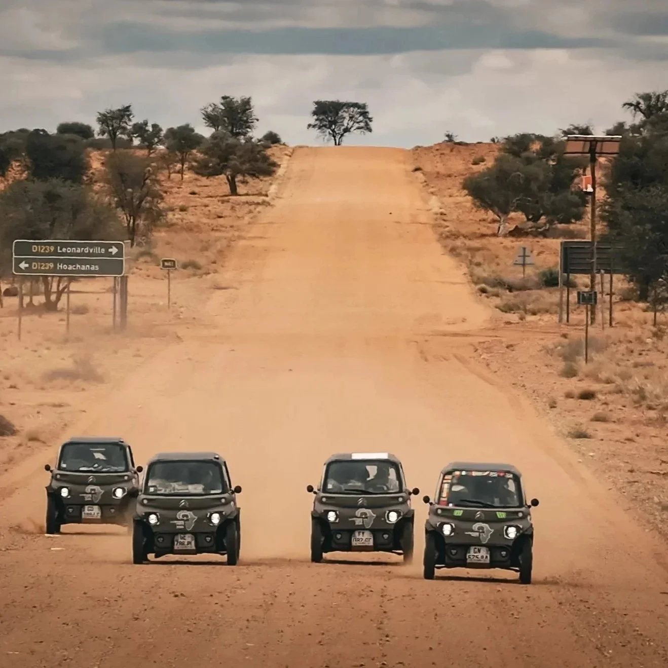 Four small black off-road vehicles parked side by side on a desert road with a steep dirt incline ahead. The landscape is dry with sparse trees and bushes, under a partly cloudy sky. Road signs indicate directions to Leonardville and Hoachanias.