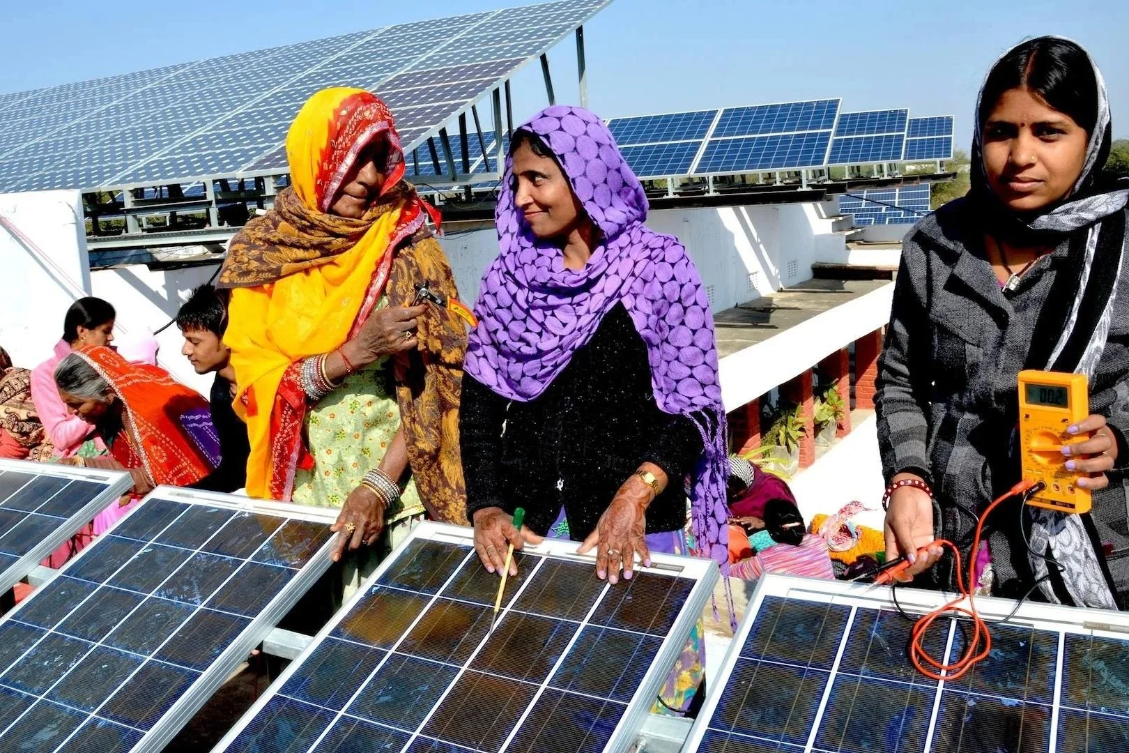 Group of women and men working on solar panels on a rooftop, with solar arrays in the background.