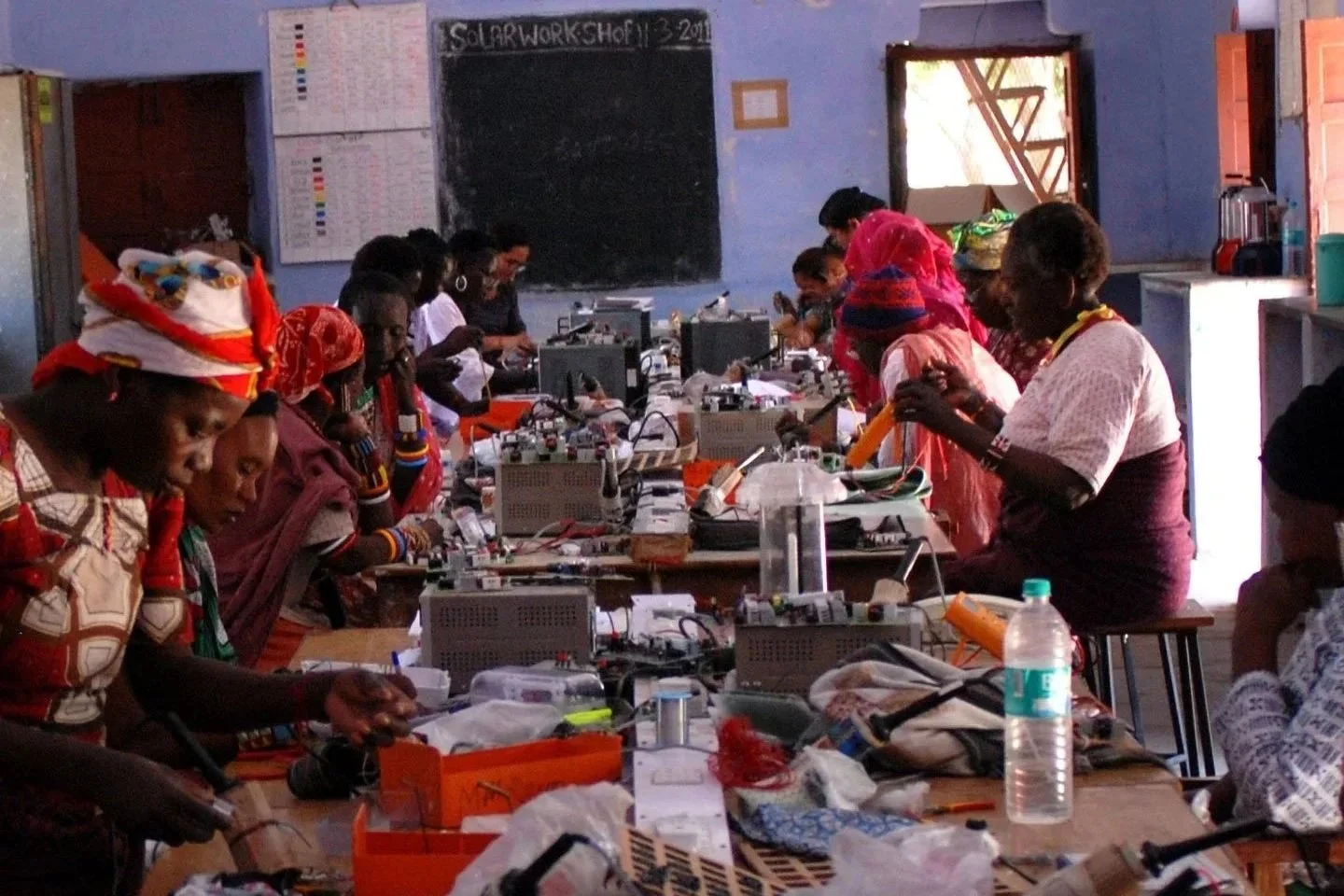 Women working on electronics projects at a table in a classroom with a blackboard and posters on the wall.