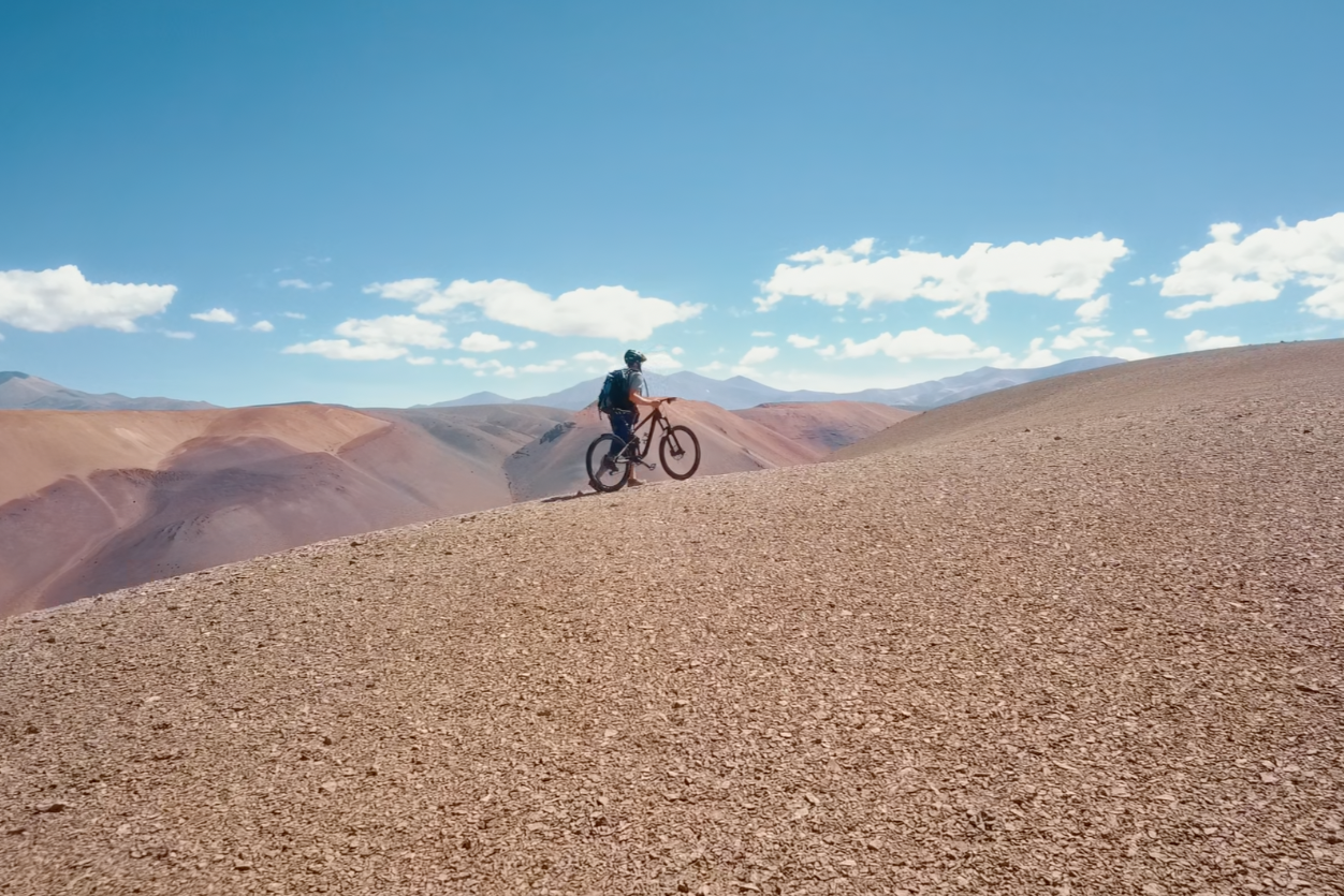 A person riding a mountain bike on a dirt trail in a desert landscape with rolling hills and mountains in the background under a blue sky with scattered clouds.