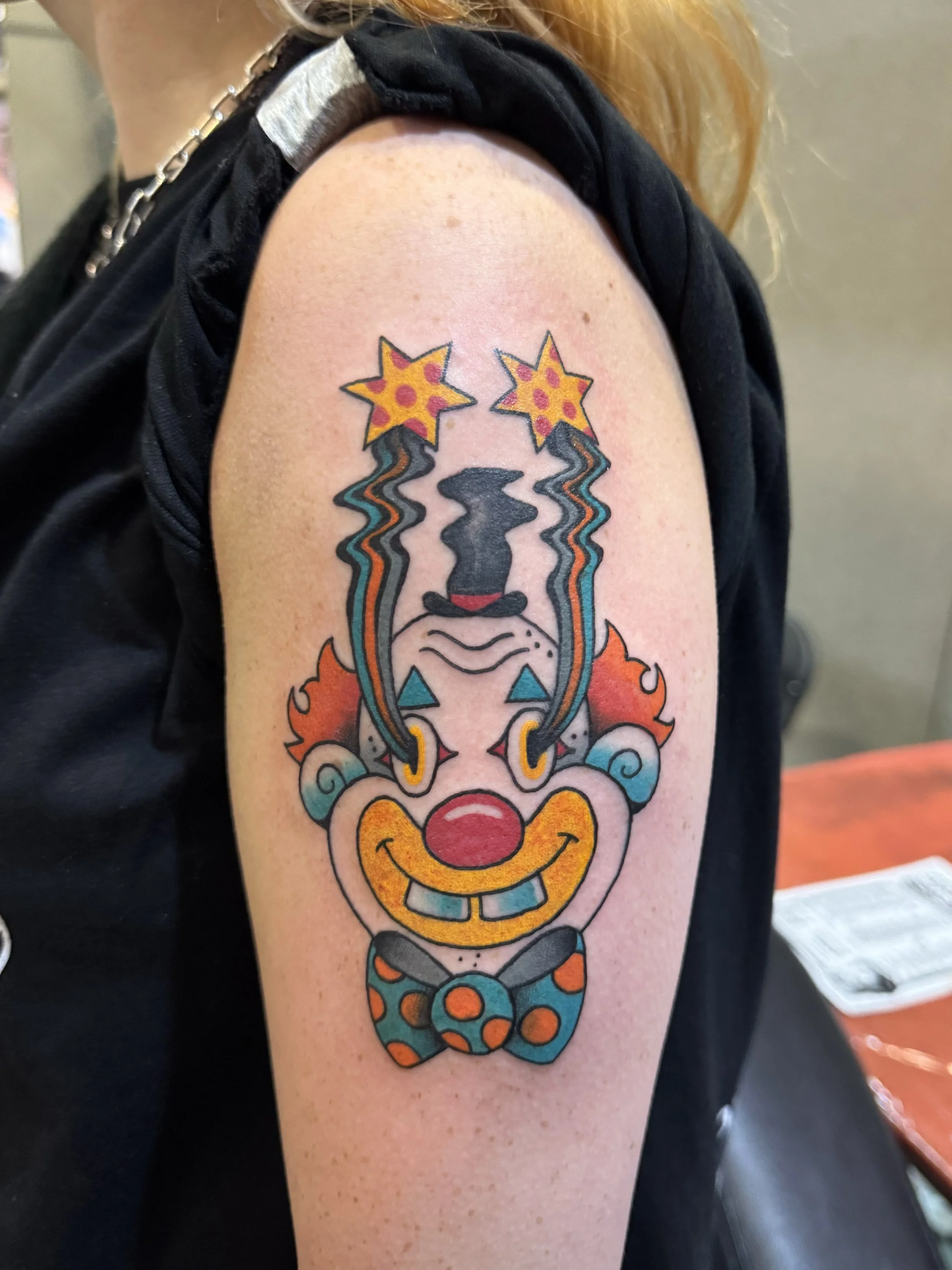 Tattoo of a  trippy clown face with orange curly hair, red nose, big yellow smile, blue makeup, wearing a blue polka dot bow tie, with two wavy lines ending in shooting star shapes above its head.