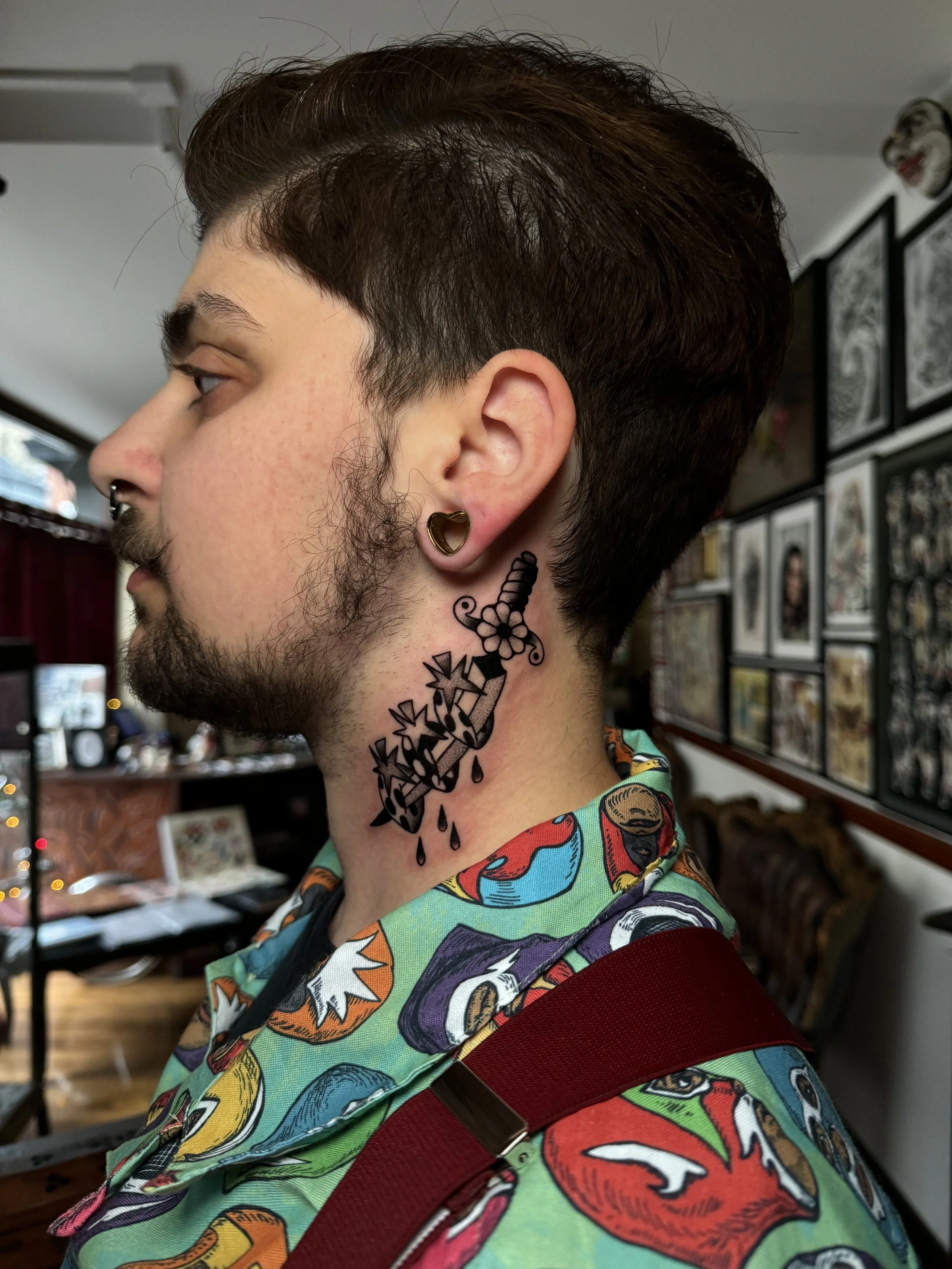 Profile of a young man with dark hair, facial hair, and multiple tattoos on his neck, including a dagger, flowers, and droplets. He has an earring in his ear, and is wearing a colorful shirt with cartoon character prints. Behind him are framed artwor