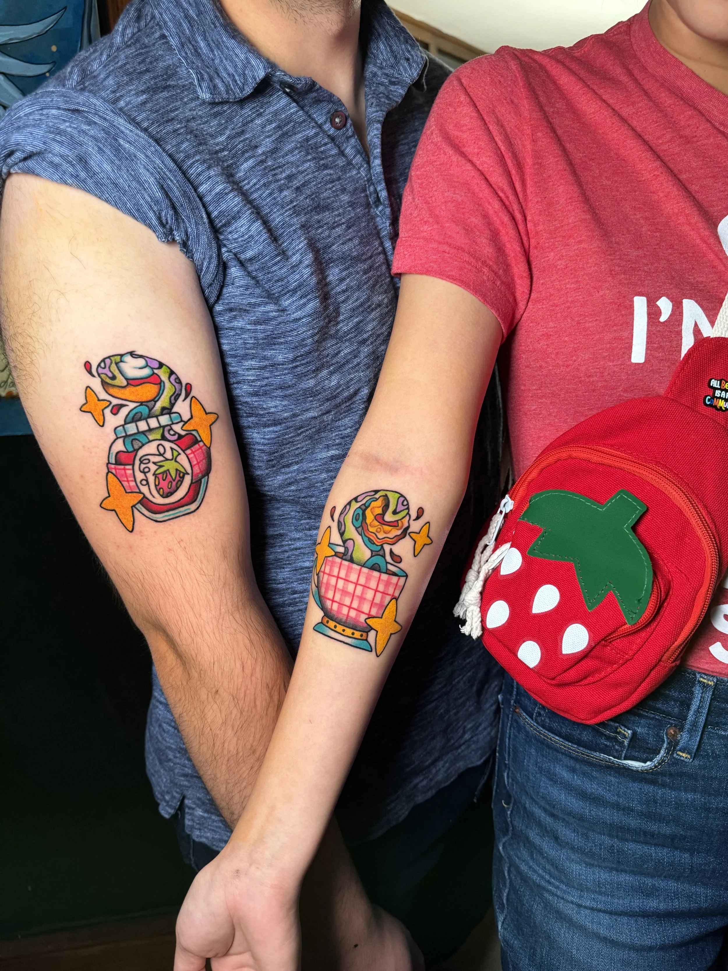 Two people showing their colorful tattoos of britsh tea and food with tentacles emerging from them. One wears a blue shirt and the other a red shirt. There is a strawberry-shaped bag near the person in red.