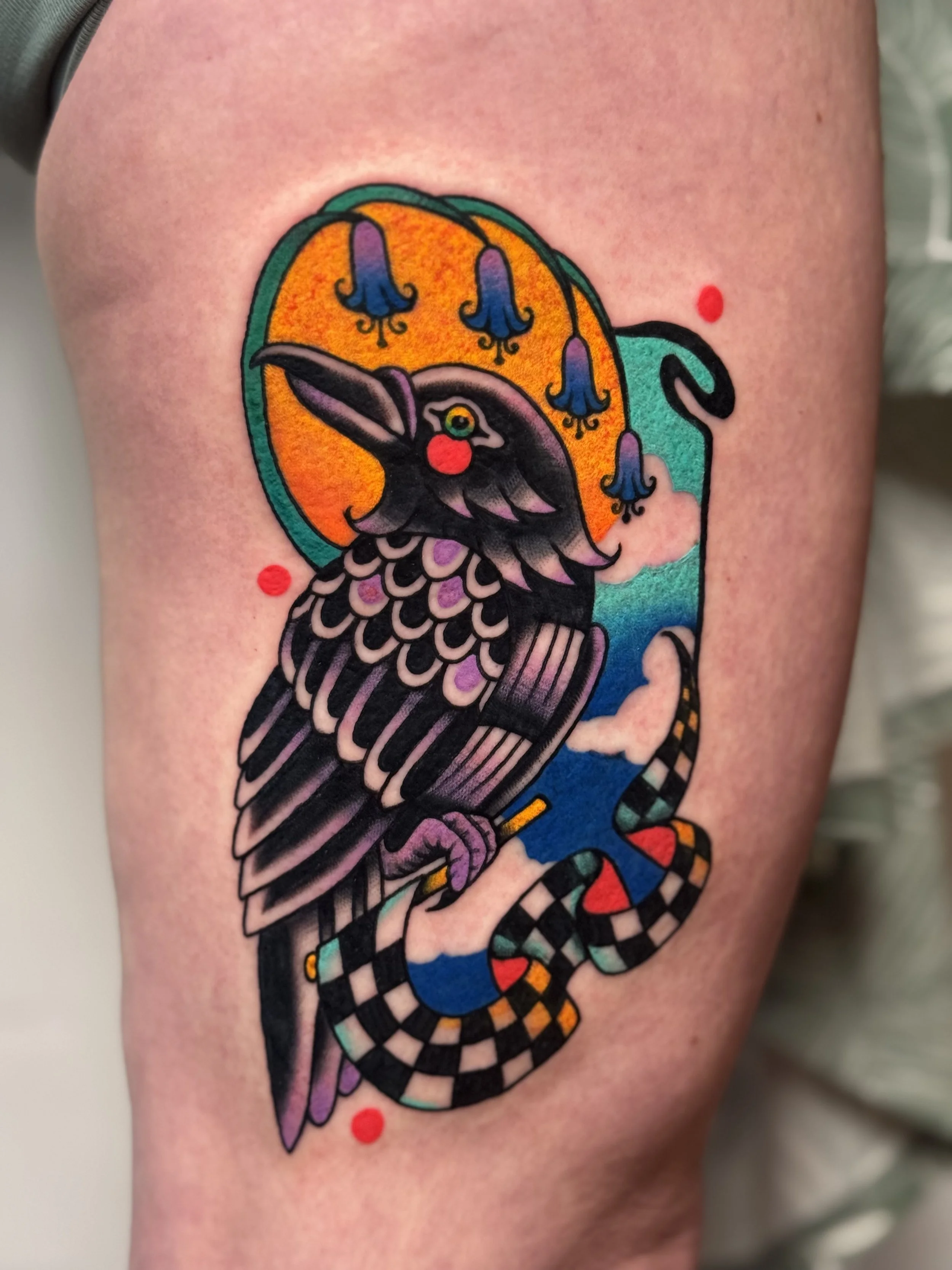 Colorful tattoo of a bird with detailed feathers, surrounded by vibrant abstract background including a yellow circle, blue flowers, and a black and white checkered snake with red eyes.