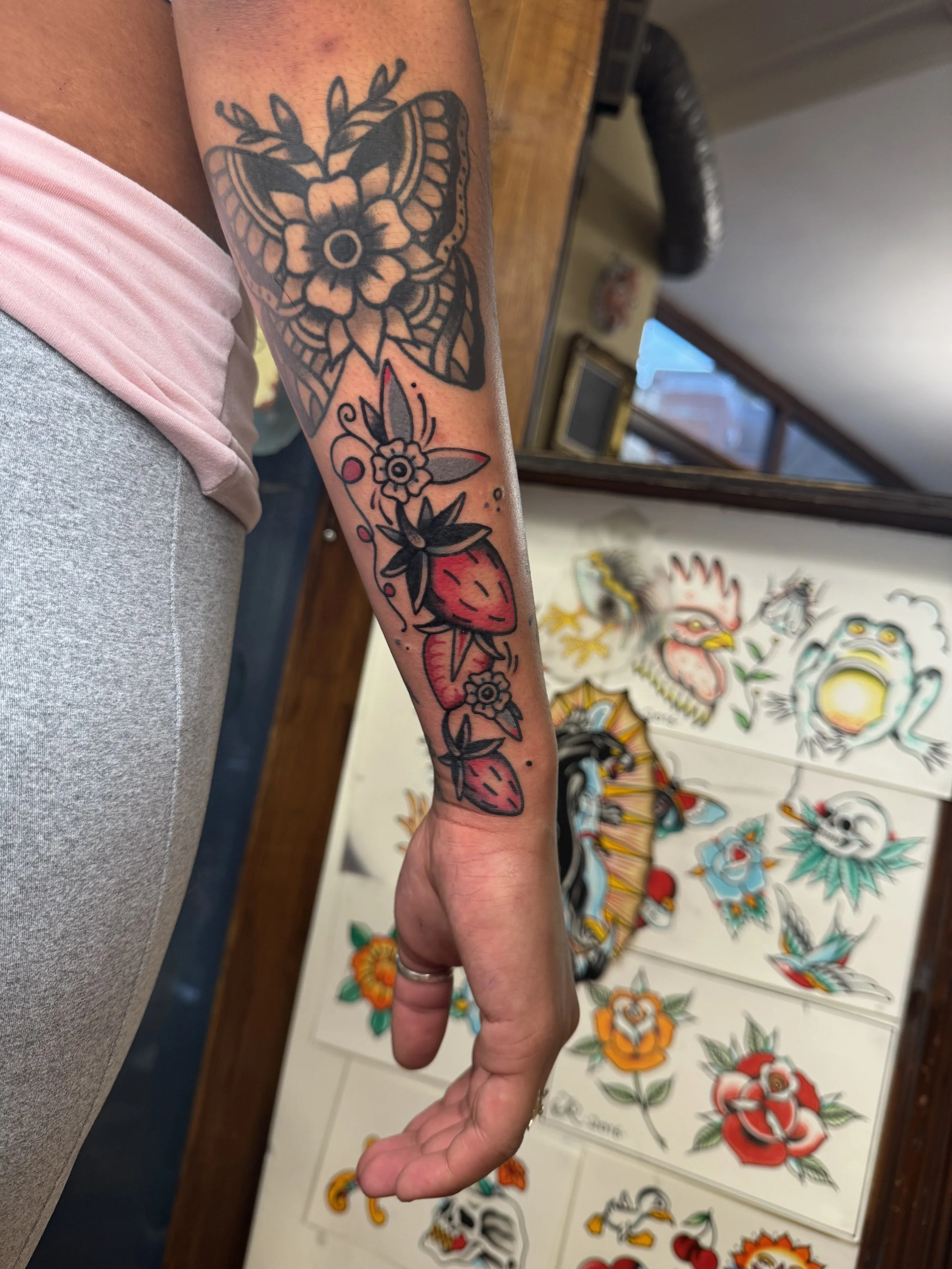 A person's arm with new tattoo of strawberries, flowers, and leaves, with artwork and sketches of traditional tattoo designs on the wall in the background.