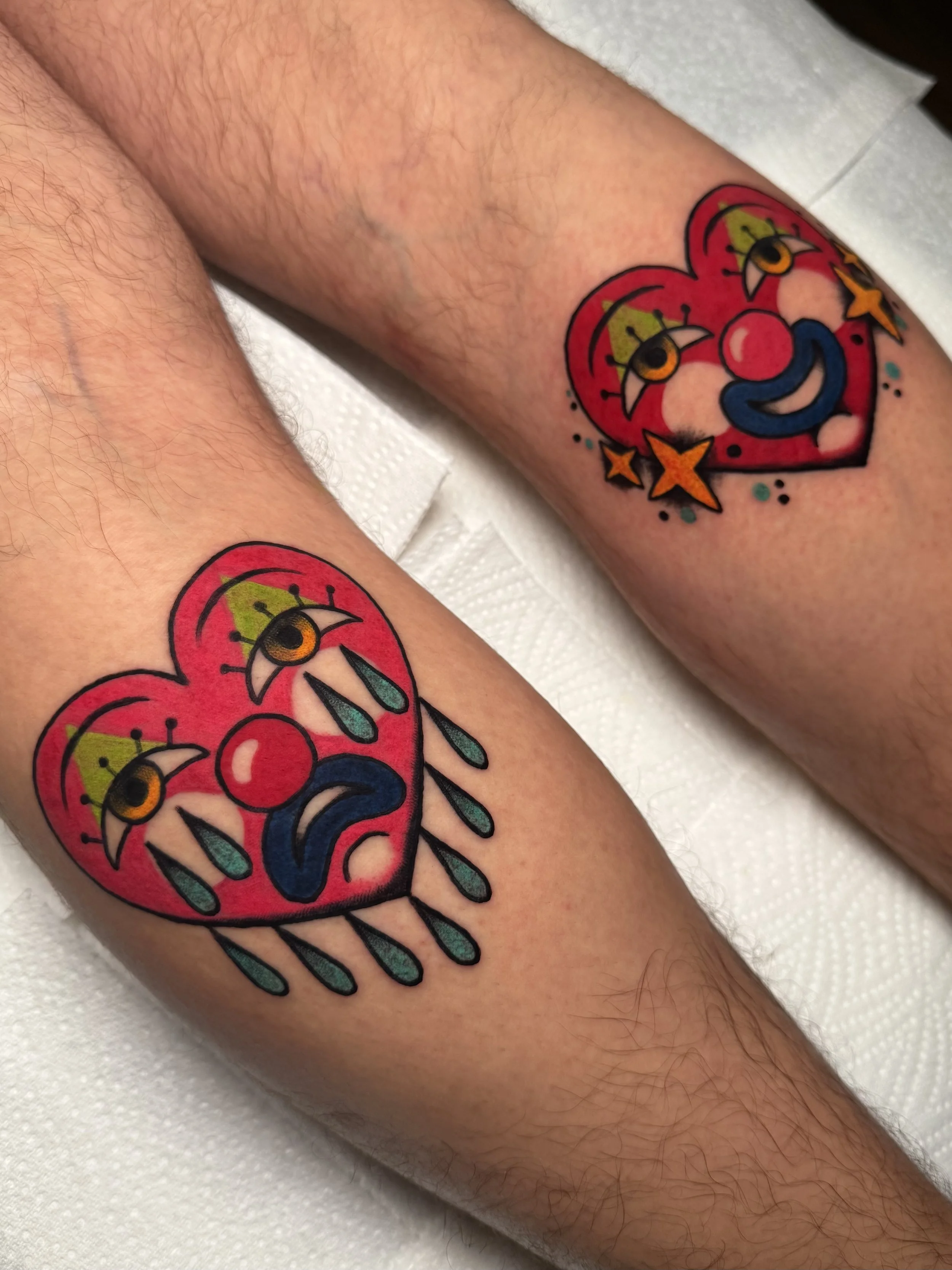 Colorful tattoo of a sad clown face inside a broken heart with teardrops, stars, and dots on a person's upper arm.