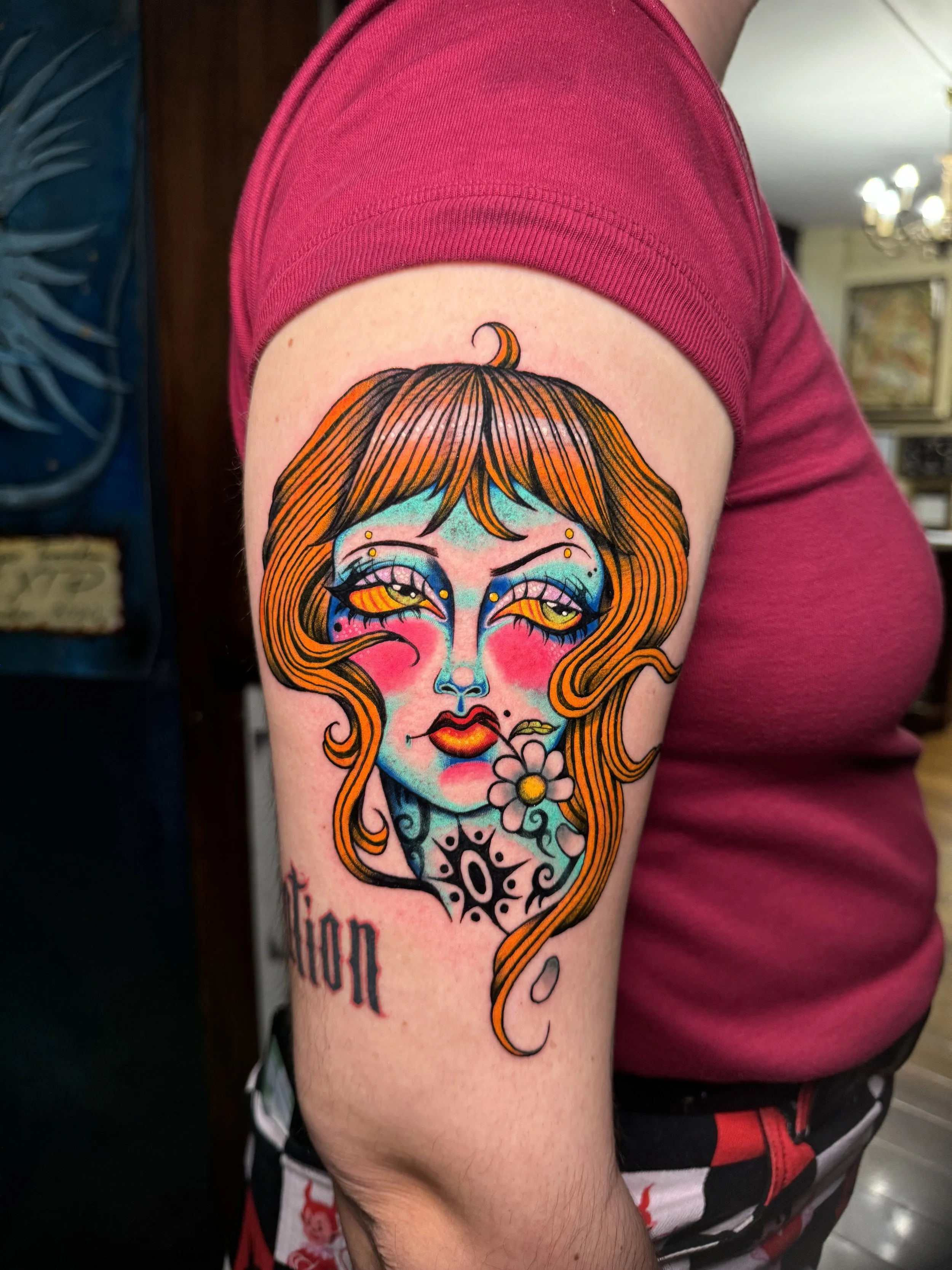 Colorful tattoo on person's upper arm depicting a woman's face with long orange hair, blue eyeshadow, pink cheeks, red lips, and a flower tattoo near her neck.