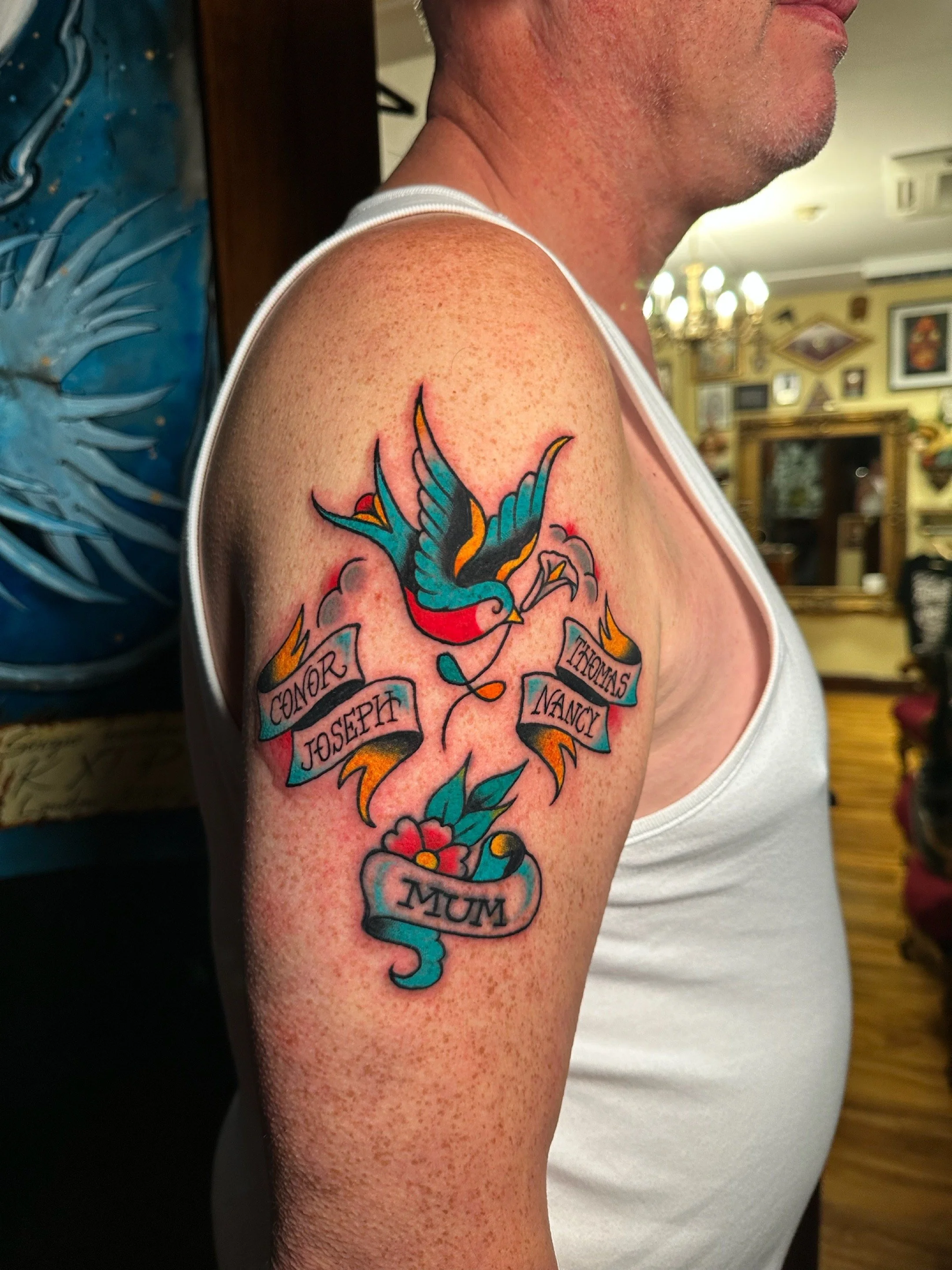 Colored tattoo of a swallow bird with a banner displaying the names Connor, Joseph, Nancy, and Synthia, and a heart-shaped ribbon with the word Mum, on a person's upper arm.