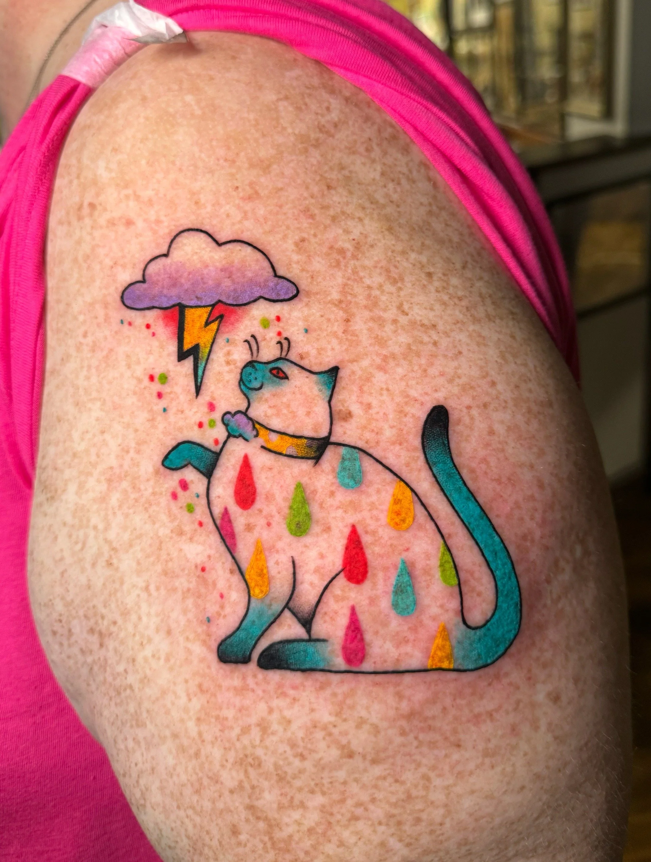 Colorful tattoo of a cat with rainbow-colored raindrops on its body, a purple cloud with a lightning bolt above its head, on a person's upper arm.