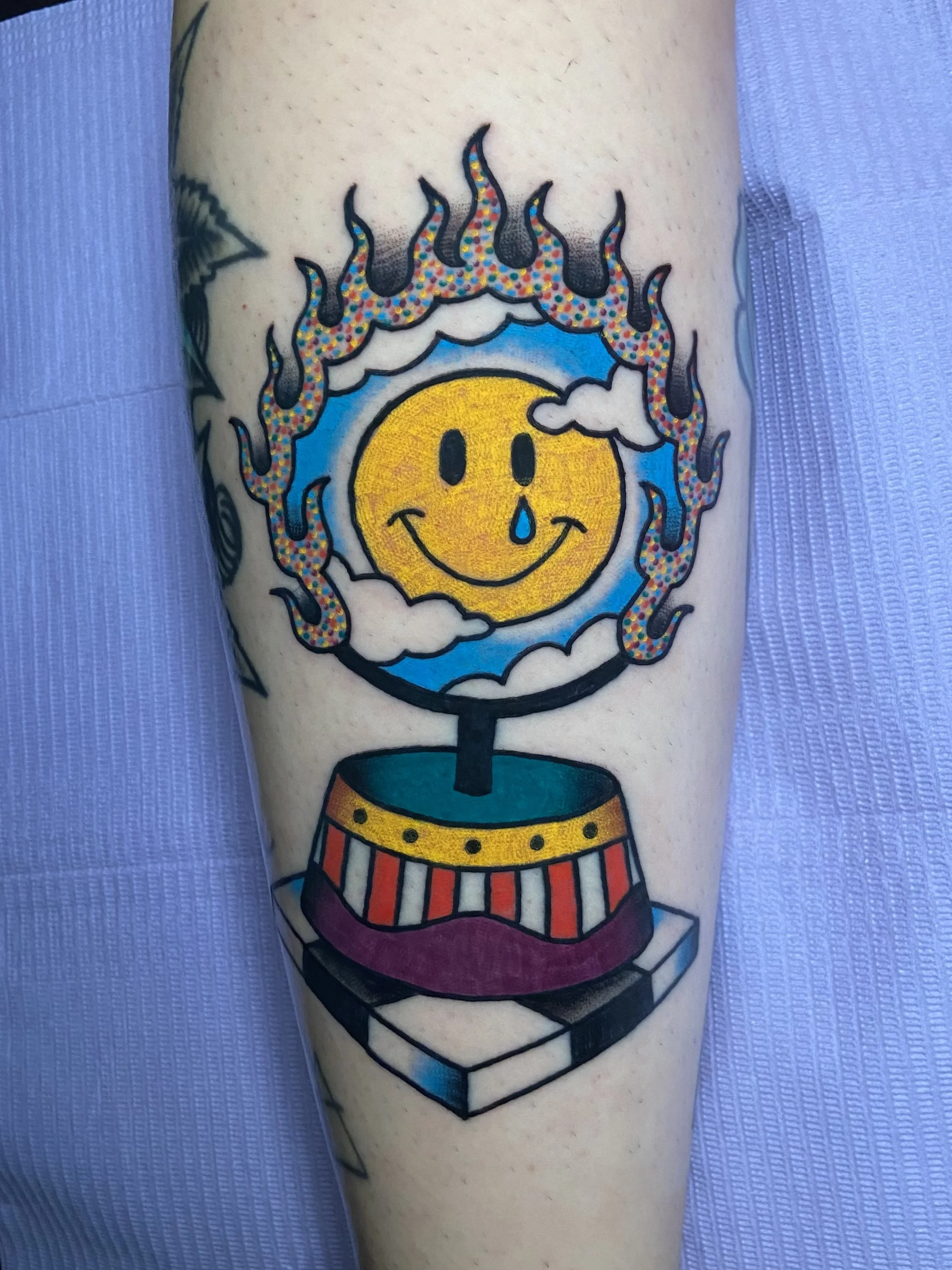 Colorful tattoo of a smiling yellow face with a tear, inside a flaming circus hoop with flames and clouds, on a person's arm.