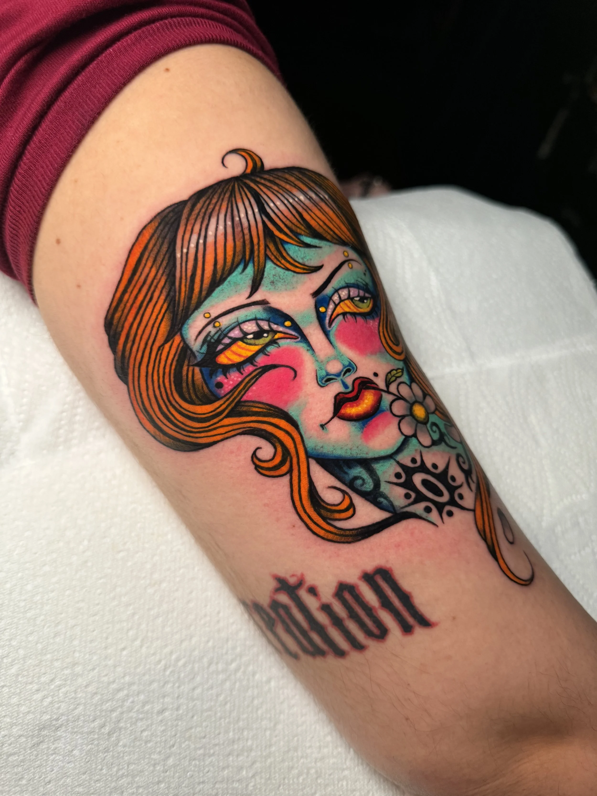 Colorful tattoo of a woman's face with orange hair, blue and pink skin tones, and yellow eyes, with a flower and text below on the upper arm.