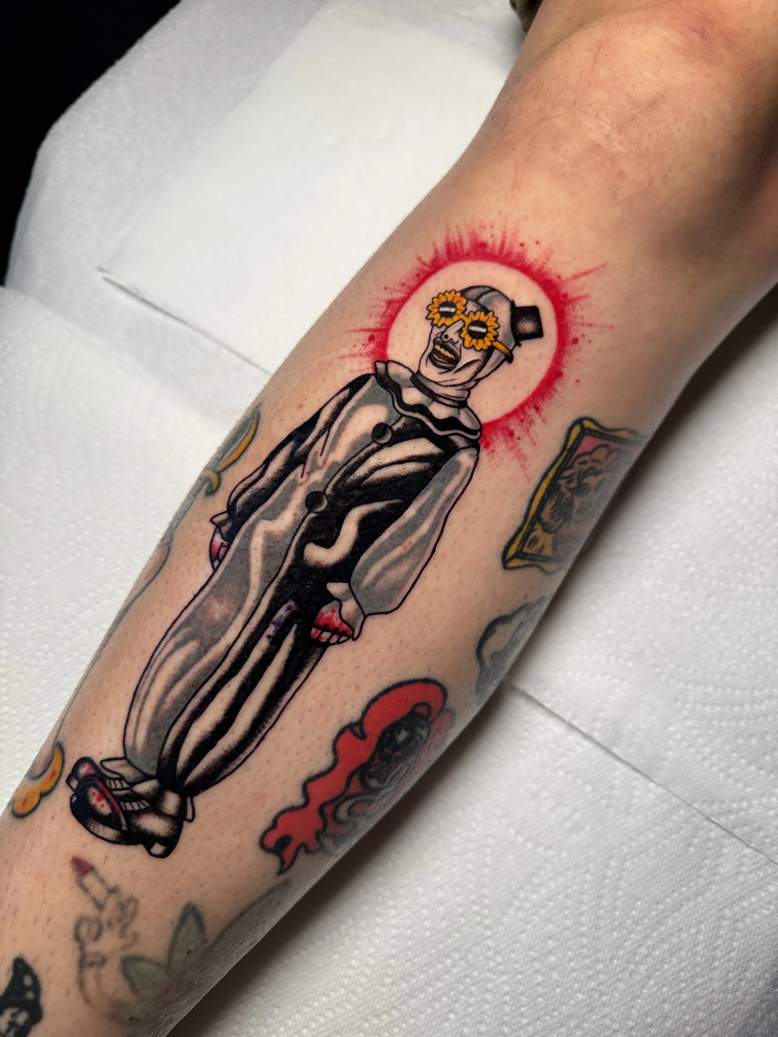 A tattoo of art the clown from the horror film the terrifier with sunflower sunglasses, wearing a black top hat and a clown outfit, surrounded by a red blood spatter halo on the arm.