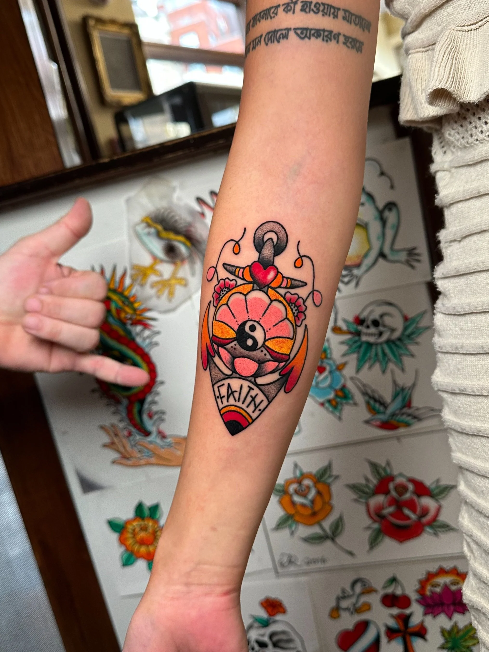 Colorful tattoo of an anchor with a yin-yang symbol in the center, featuring a heart and a banner with the word 'FAITH' beneath, on a person's forearm.