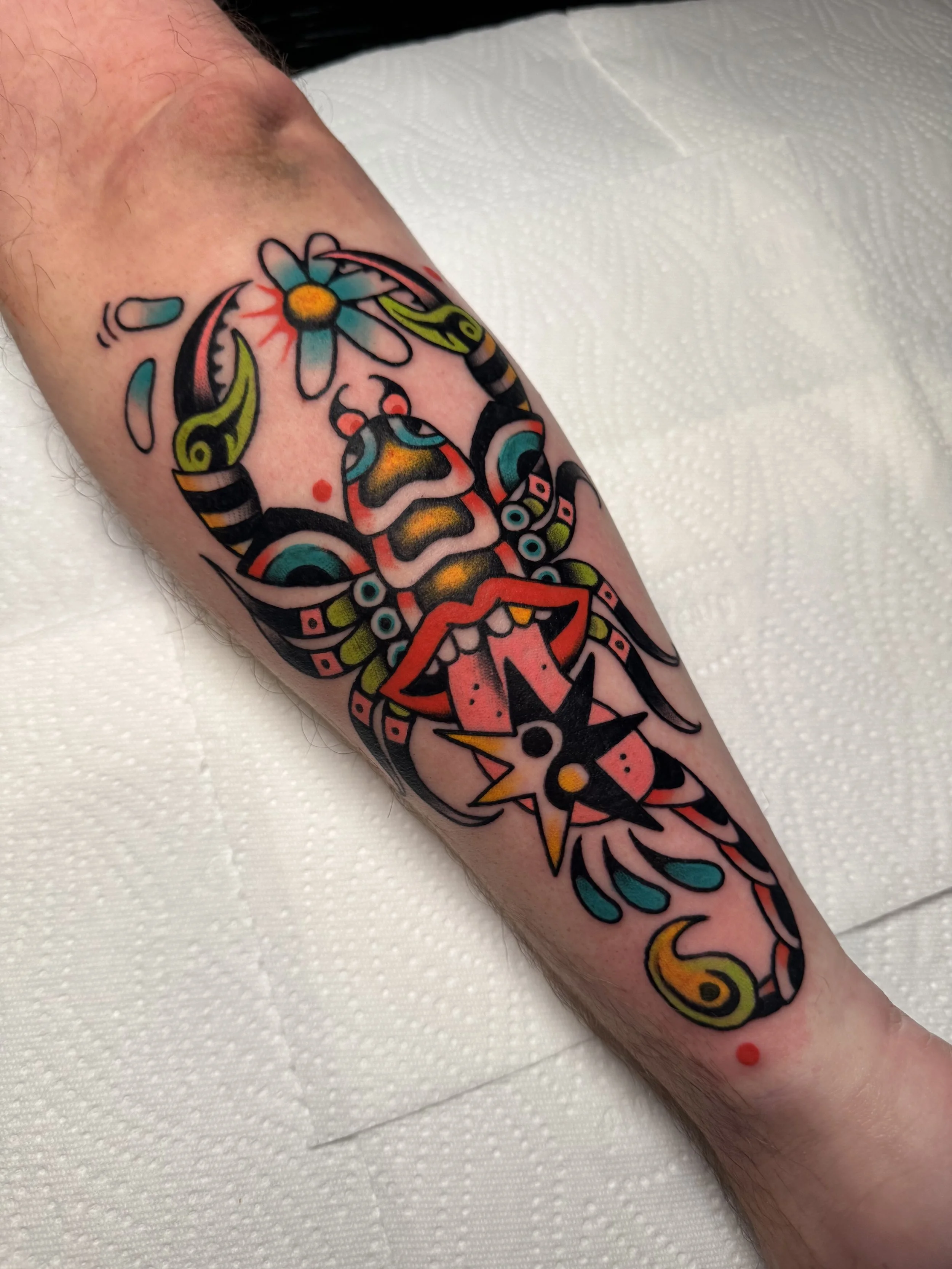 Colorful traditional tattoo of a scorpion with a flower on its back on a person's forearm, featuring bold black outlines and vibrant shades of red, yellow, green, blue, and orange.