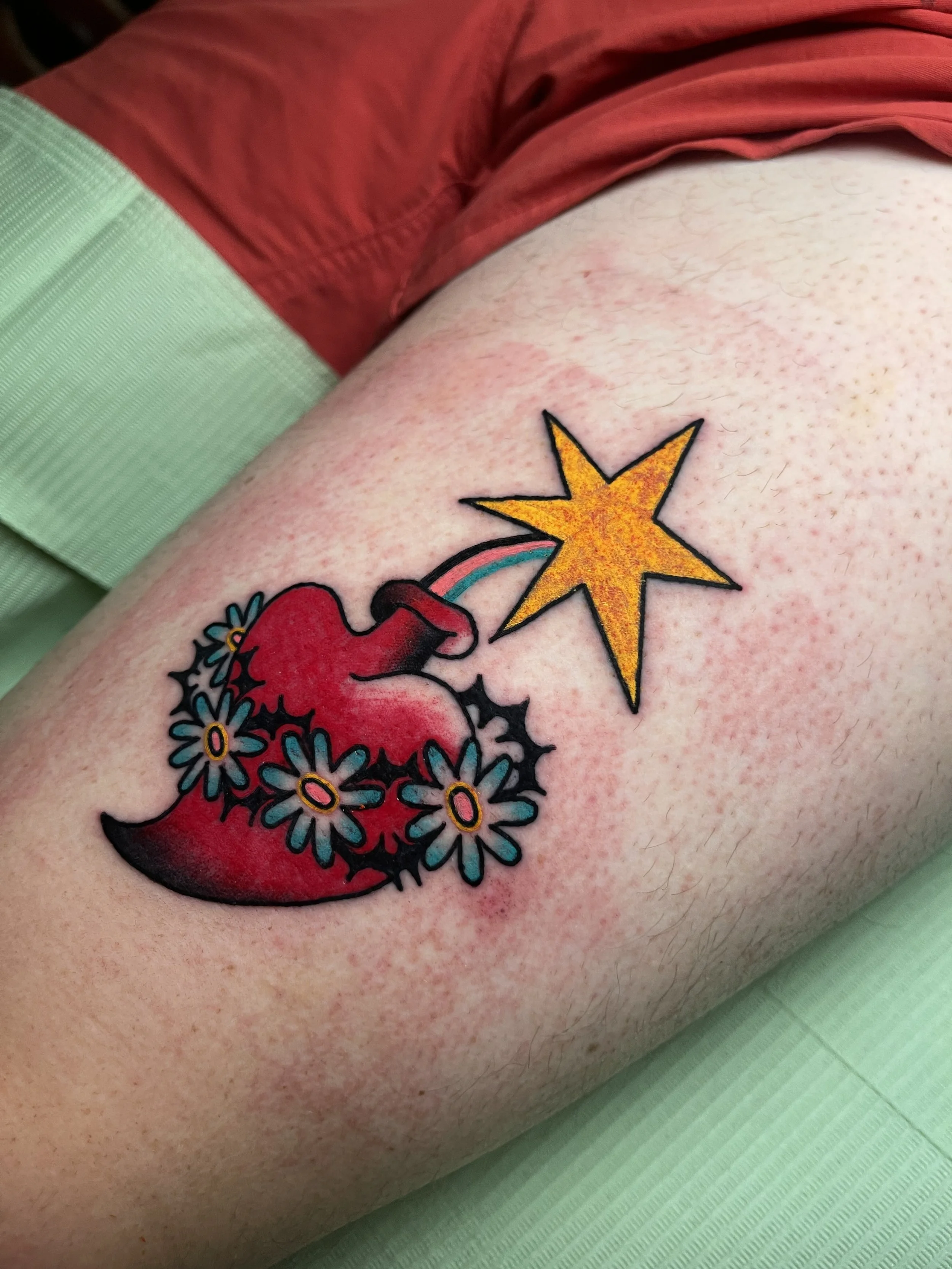 Tattoo of a red sacred heart with flowers and a yellow star on a person's thigh.
