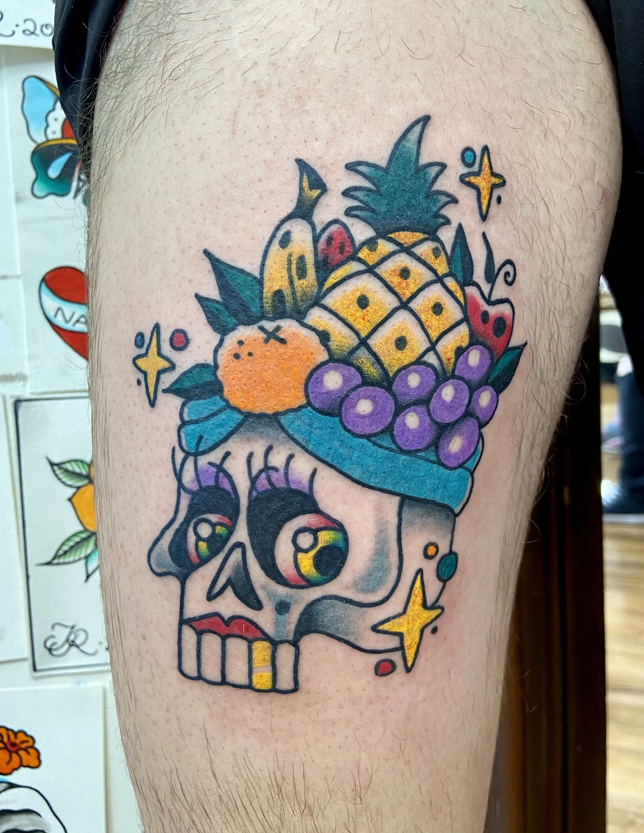 Tattoo of a skull with a fruit headdress including a pineapple, grapes, and other fruits in colorful traditional tattoo style.