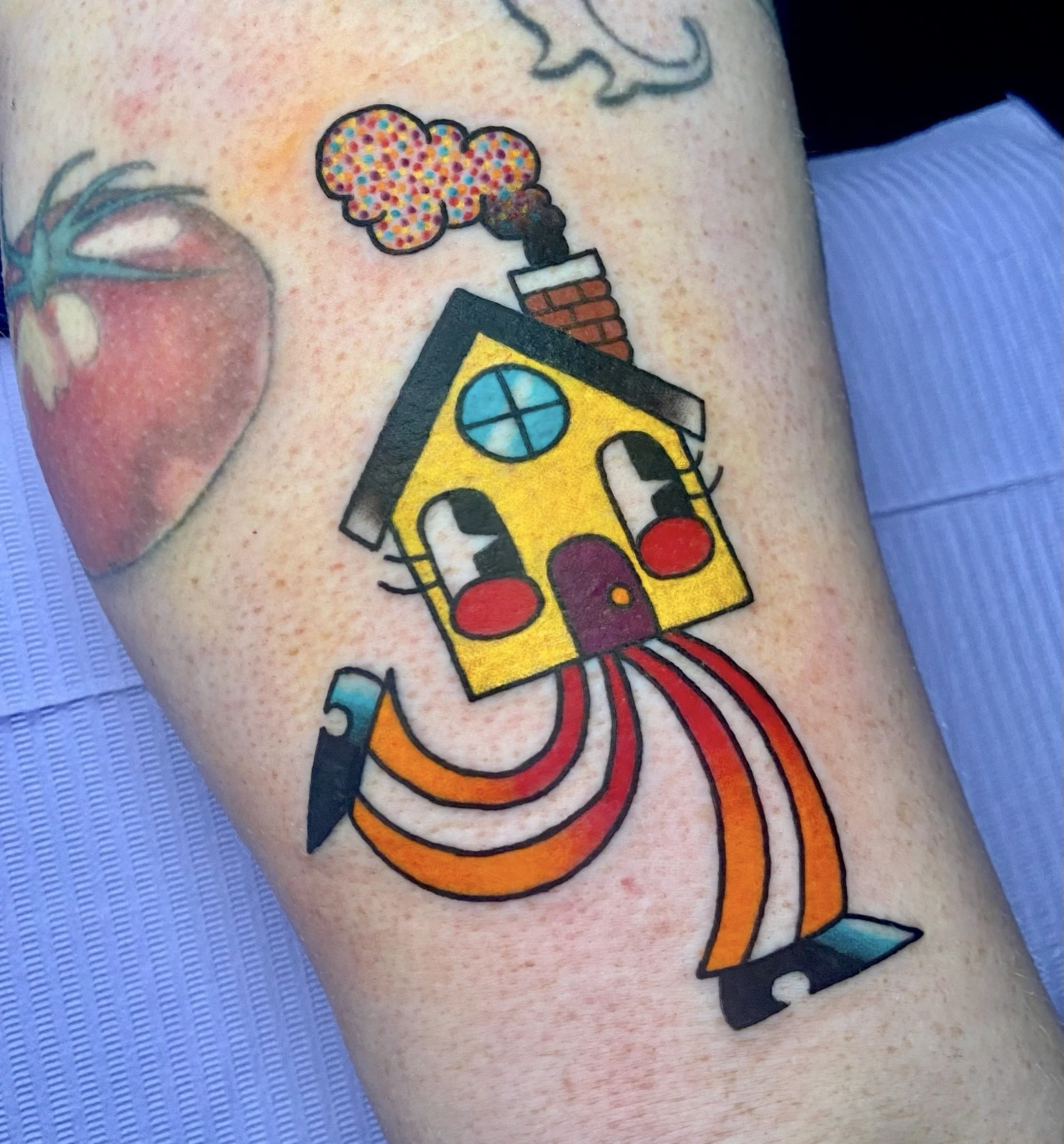 Colorful tattoo of a cartoon house with a smiling face, red cheeks, and a chimney emitting colorful smoke, on a person's arm.