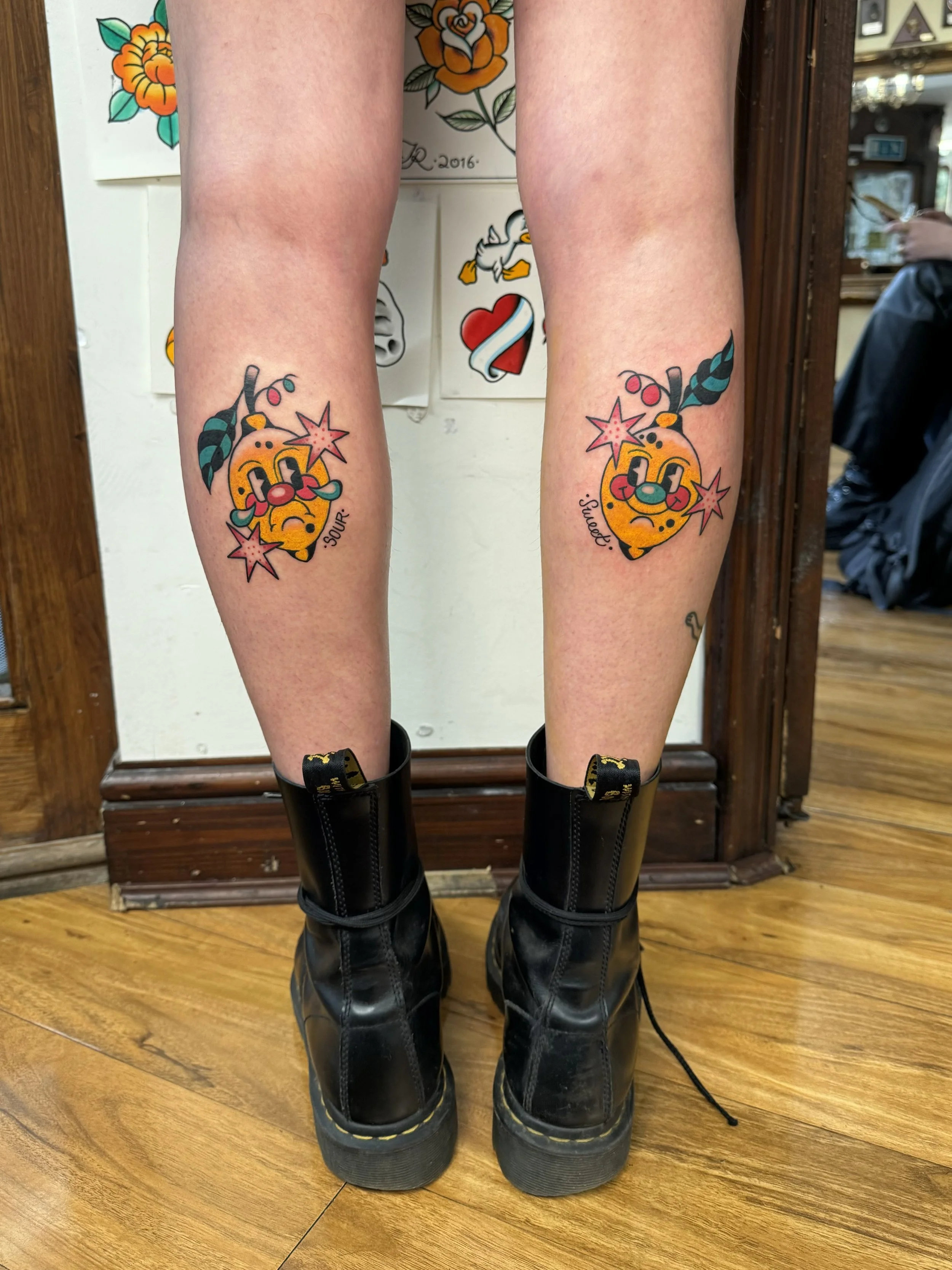 Two tattoos of cartoonish Lemons with faces surrounded by stars and cherries, on the calves of a person wearing black boots.