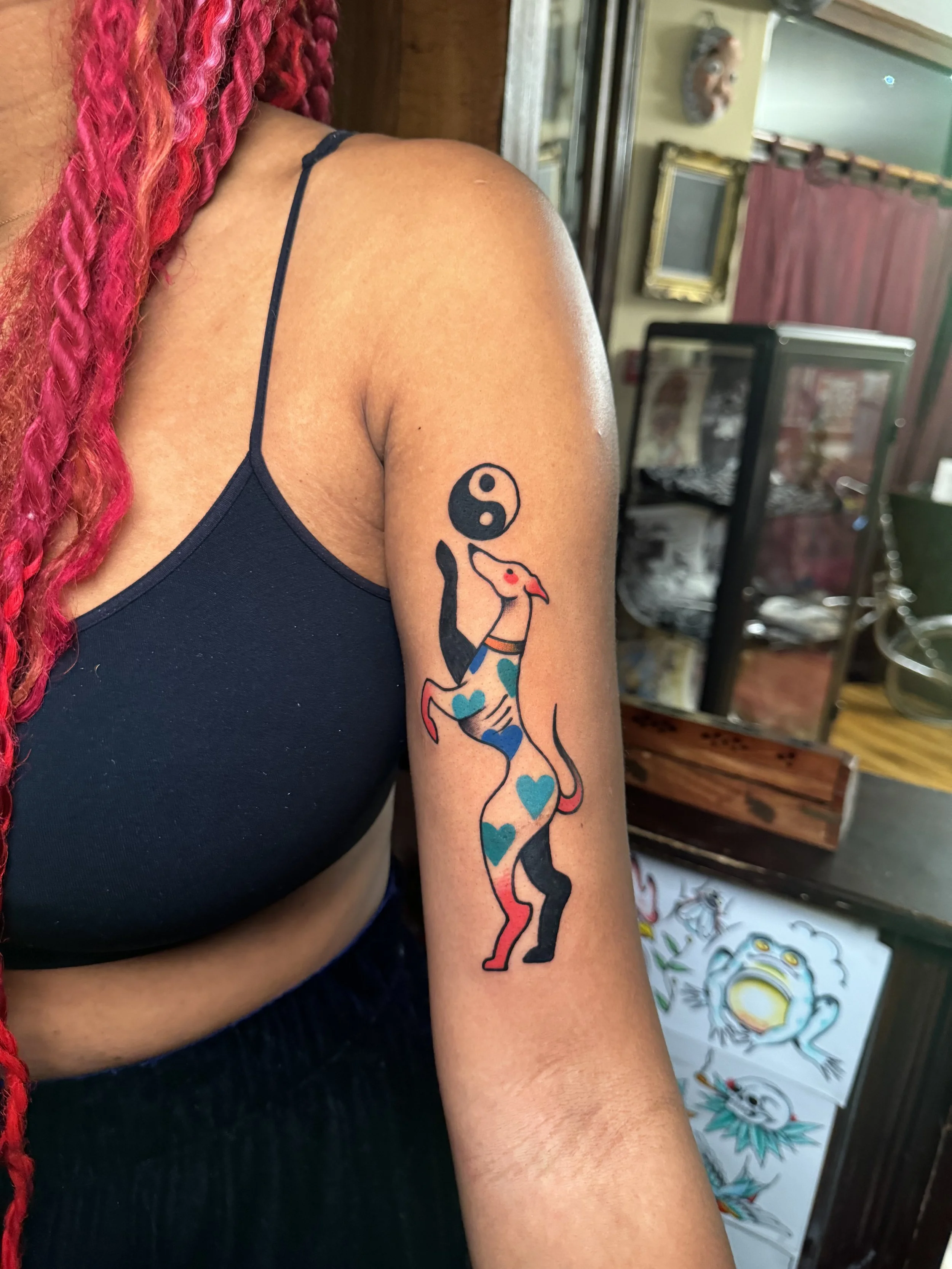 Tattoo of a dog with blue hearts on its body, holding a yin-yang symbol, on a person's upper arm.