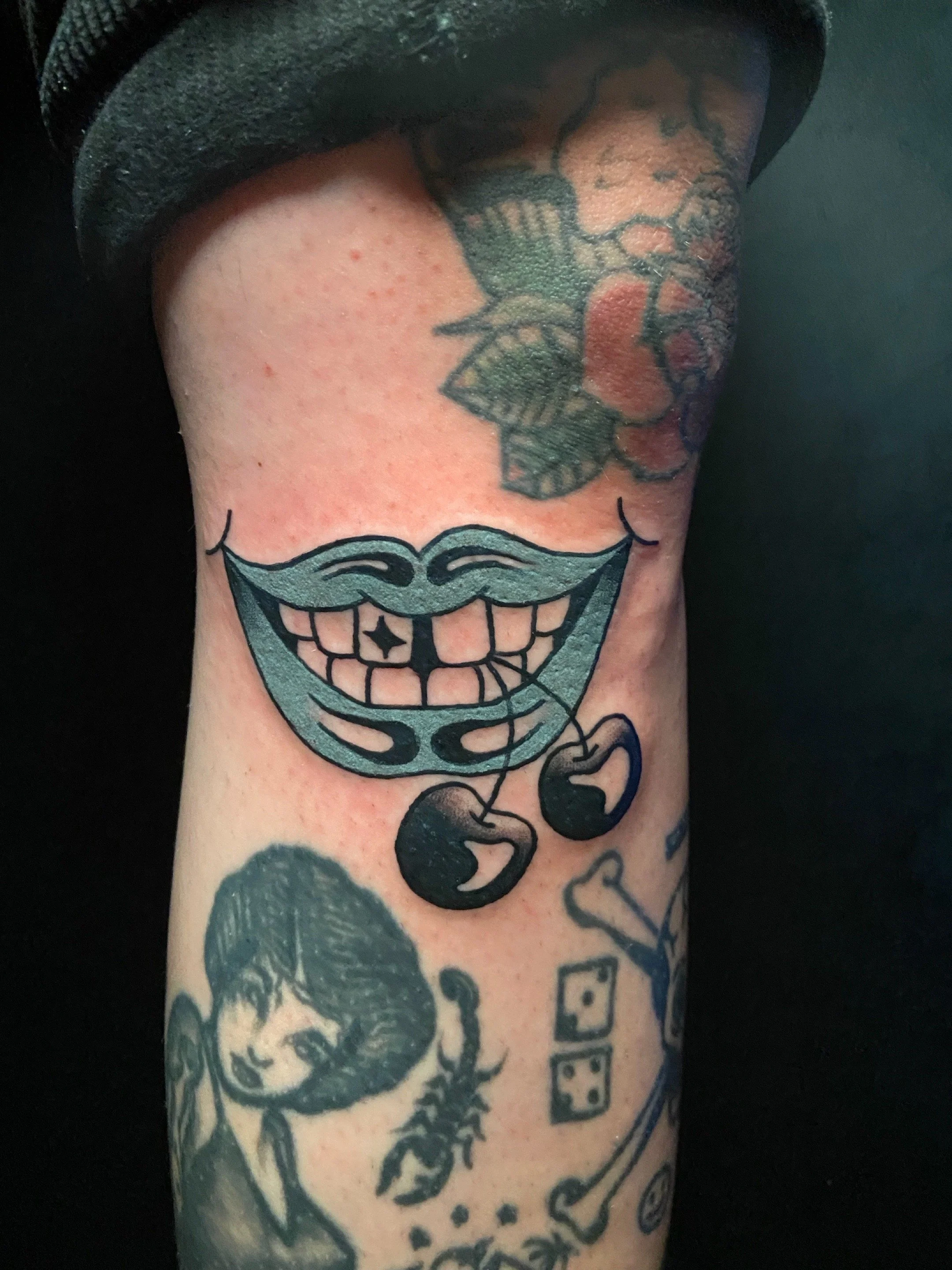 Tattoo of a smiling mouth with teeth, with two cherries hanging from the corners, on a person’s leg.