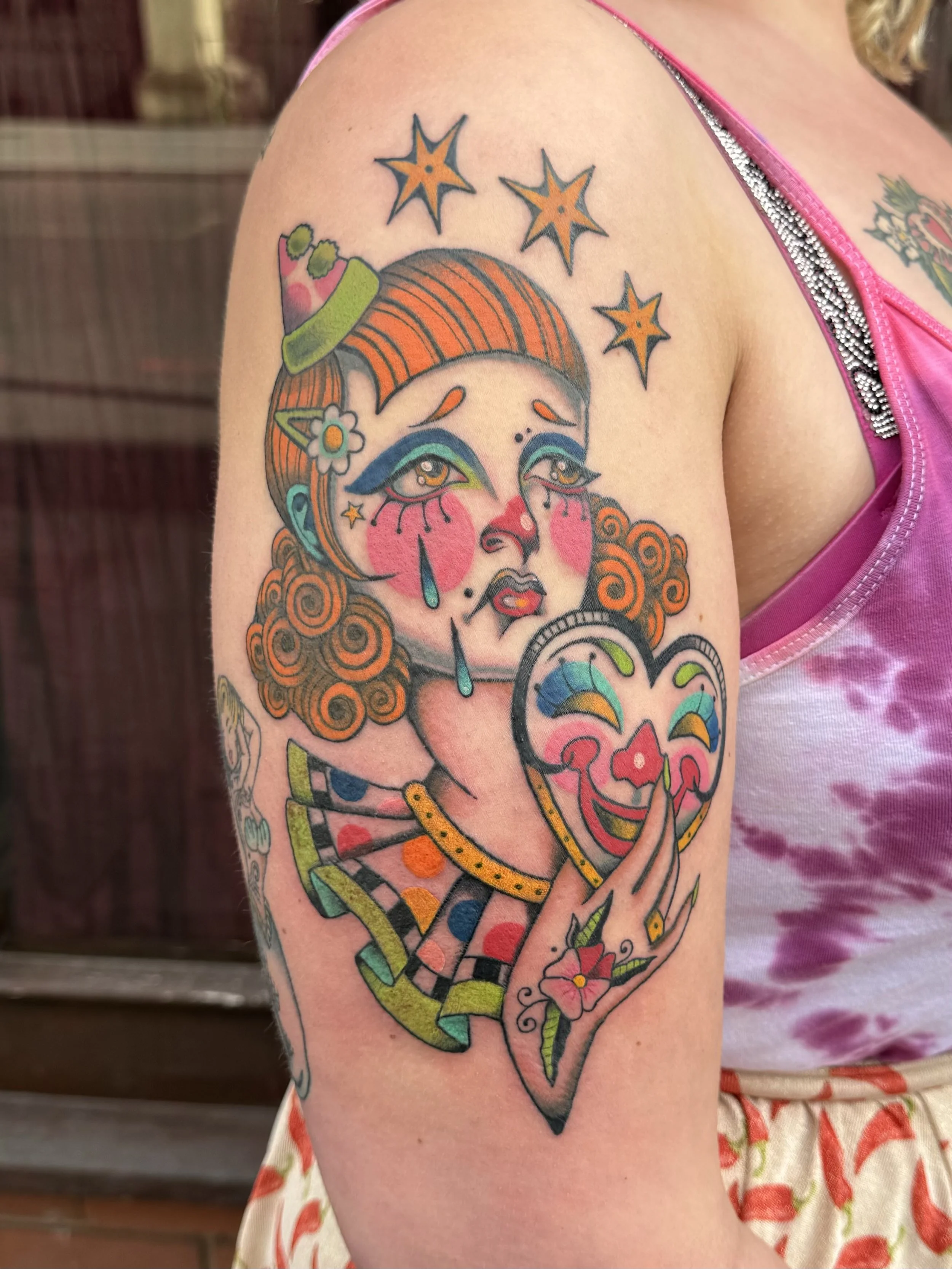 Colorful tattoo of a crying clown girl with orange curly hair, wearing a small green hat, holding a smiling clown face mask, with stars and tears around her, on a person's upper arm.