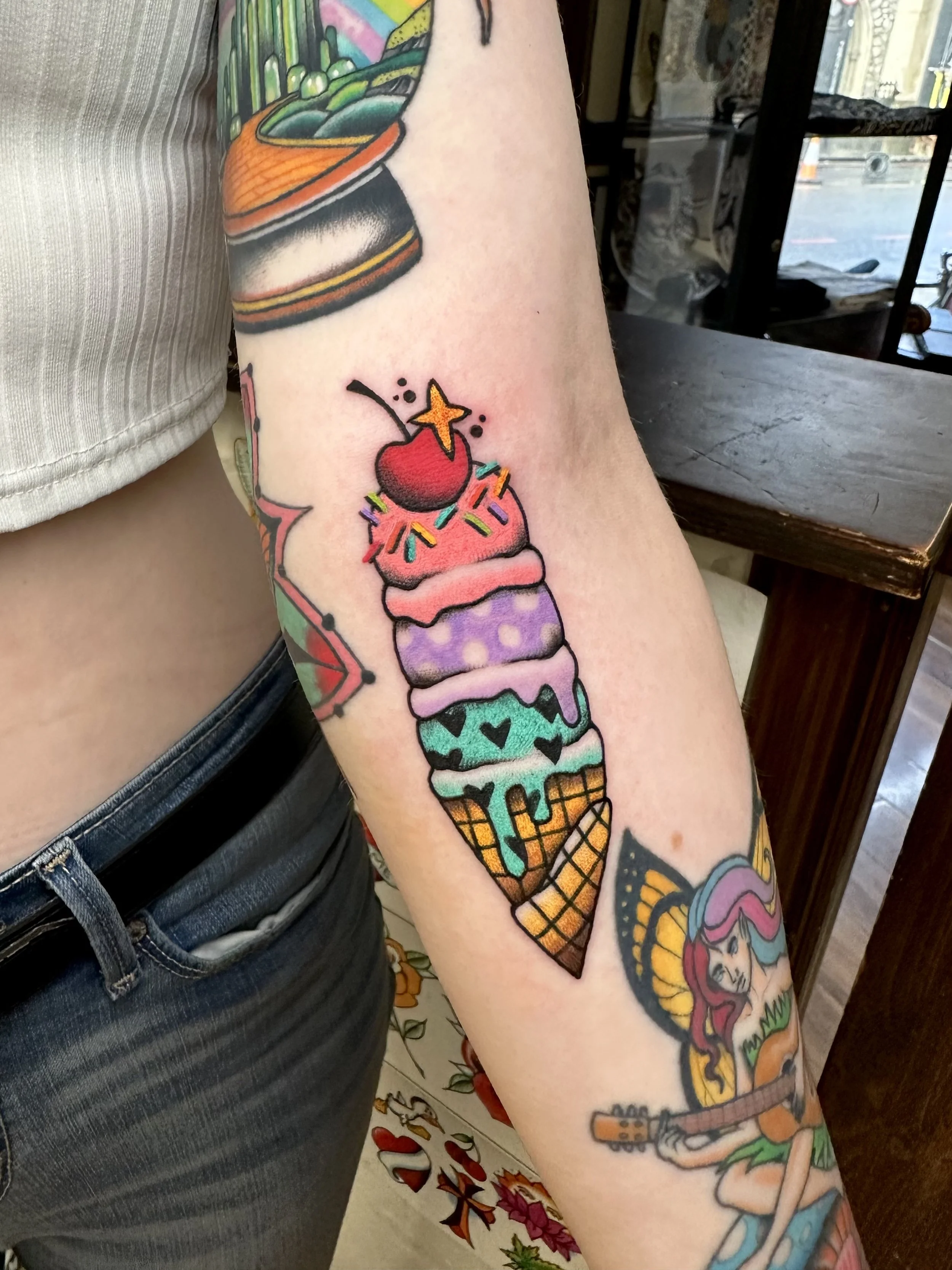 Colorful tattoo of a multi-scoop ice cream cone topped with a cherry and star-shaped decoration on a person's arm.