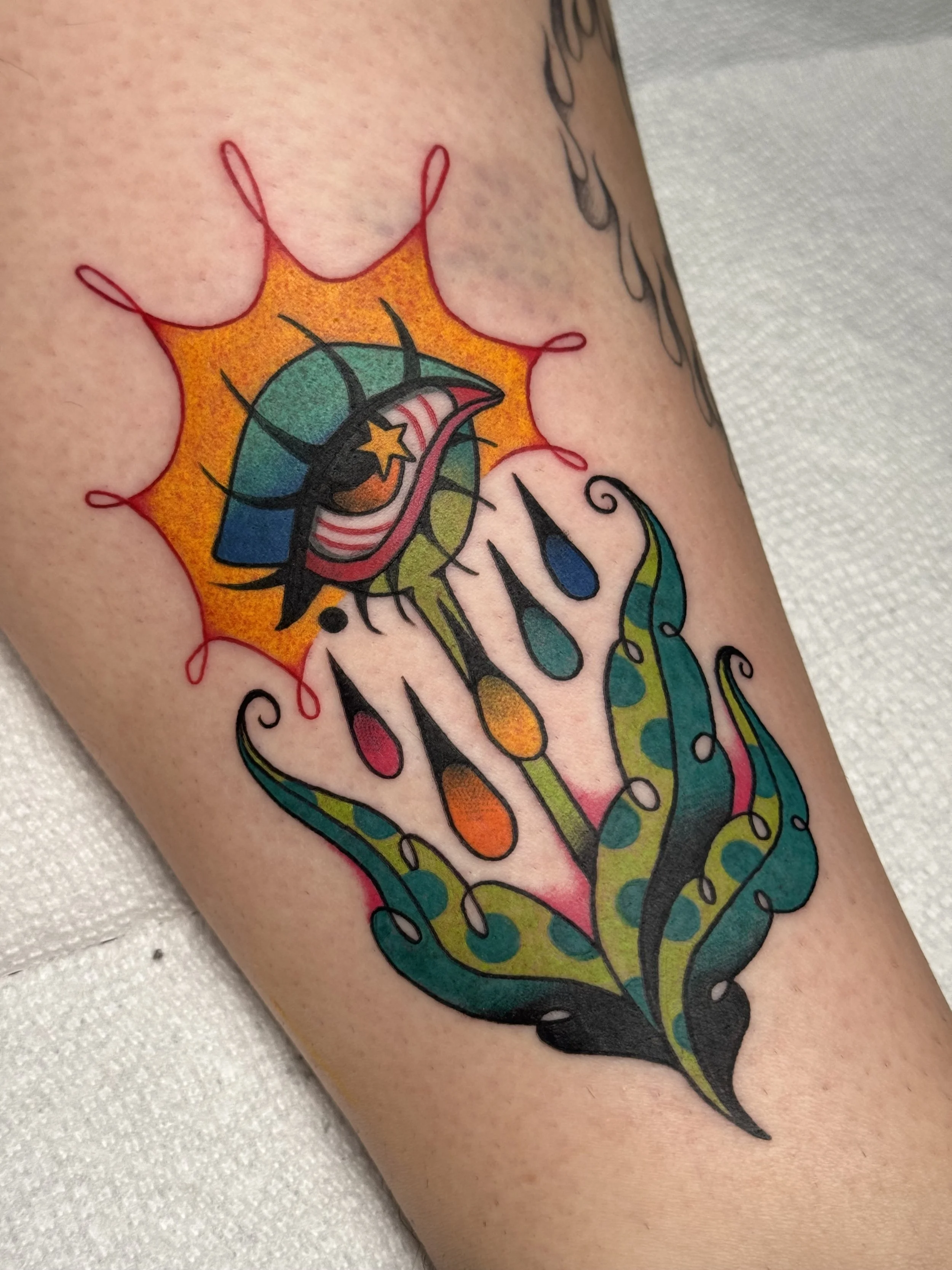 Colorful tattoo of a trippy eye flower, surrounded by an orange burst and abstract shapes in green, blue, pink, and black.