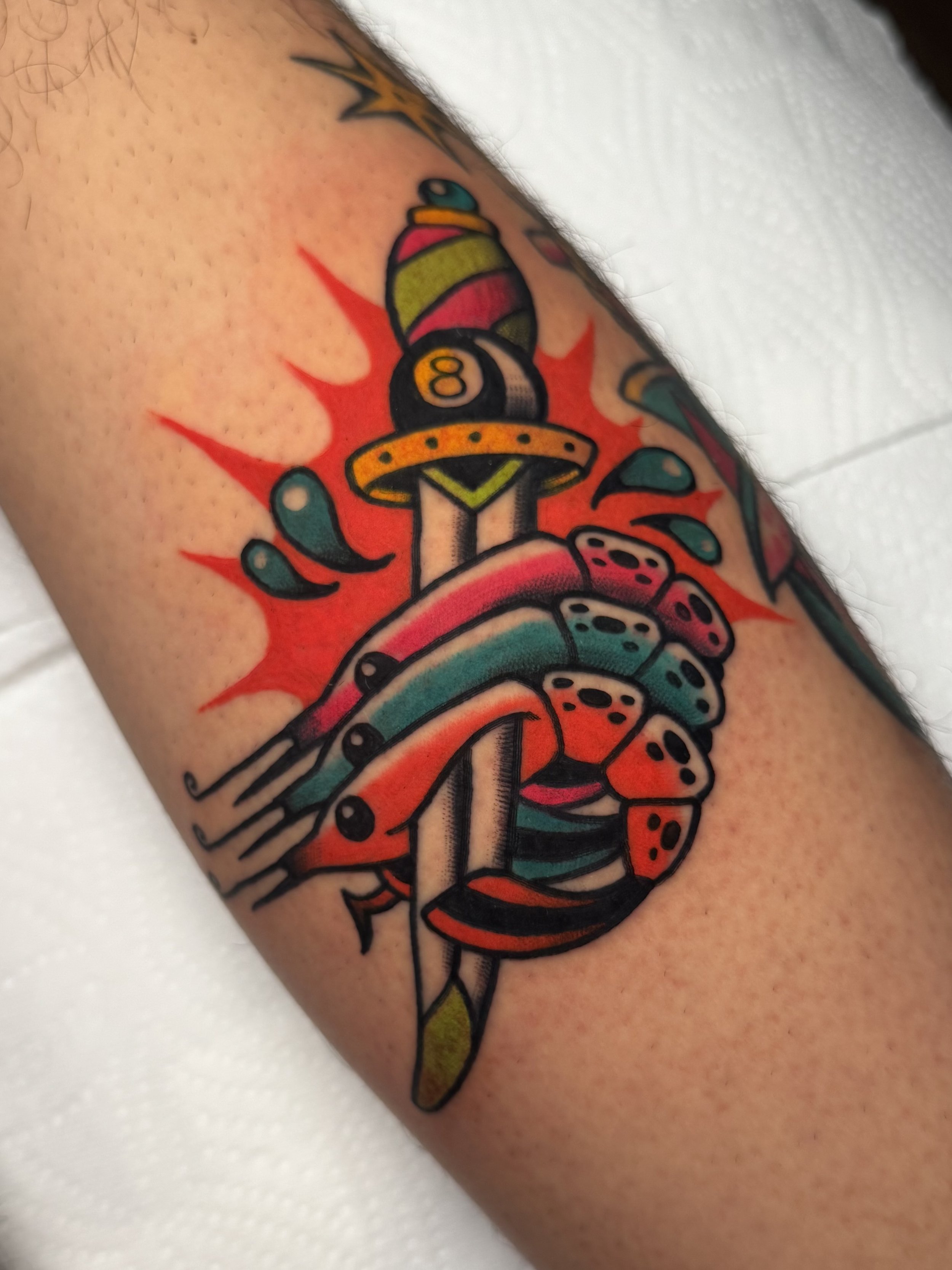 Tattoo of a dagger with a colorful, stylized hilt and blade, that has three colourful shrimp on it, surrounded by red and teal splash effects.