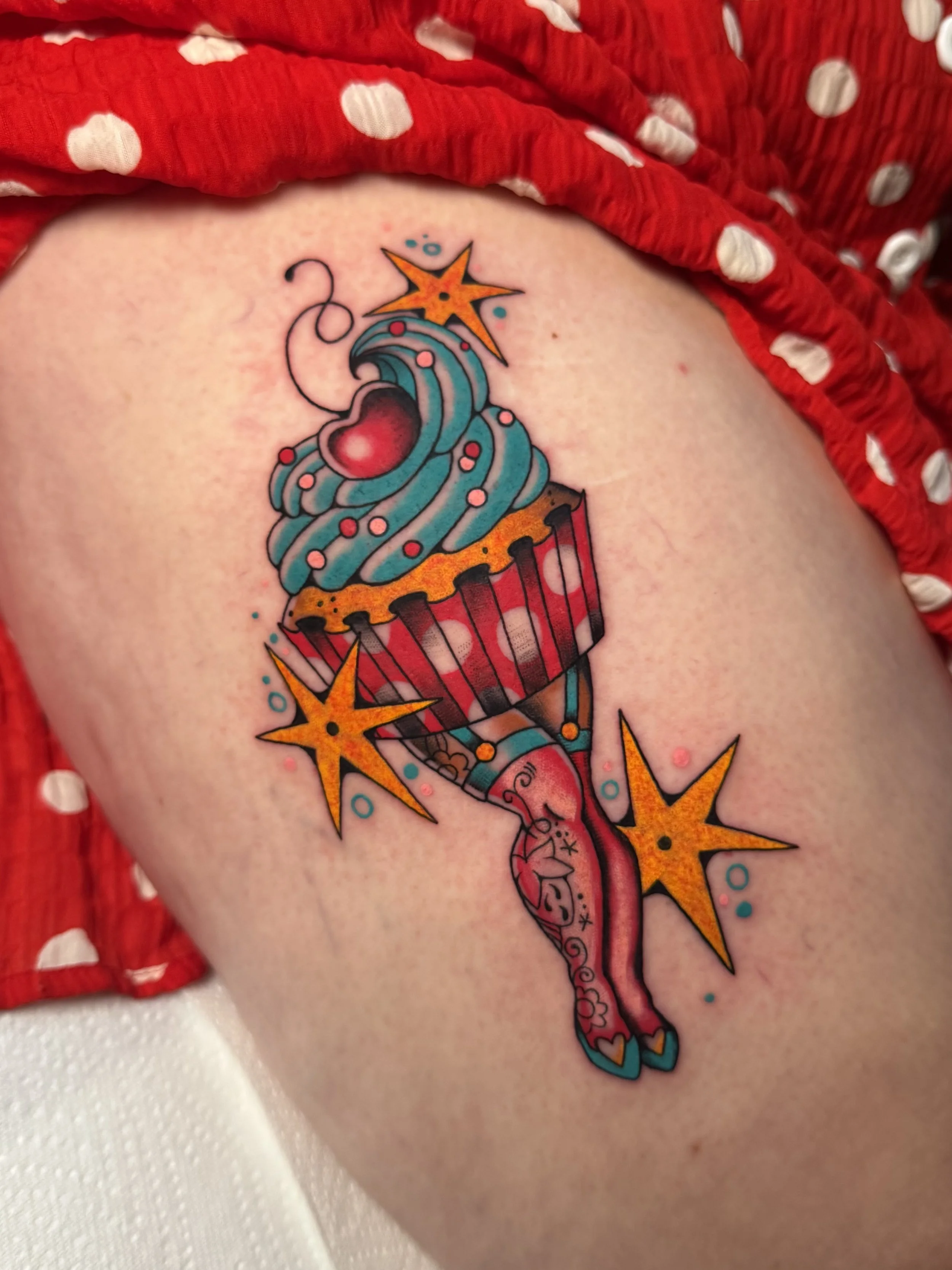 Colorful tattoo of a cupcake pin-up with blue frosting topped with a red heart and surrounded by yellow starbursts and blue dots on a person's arm.