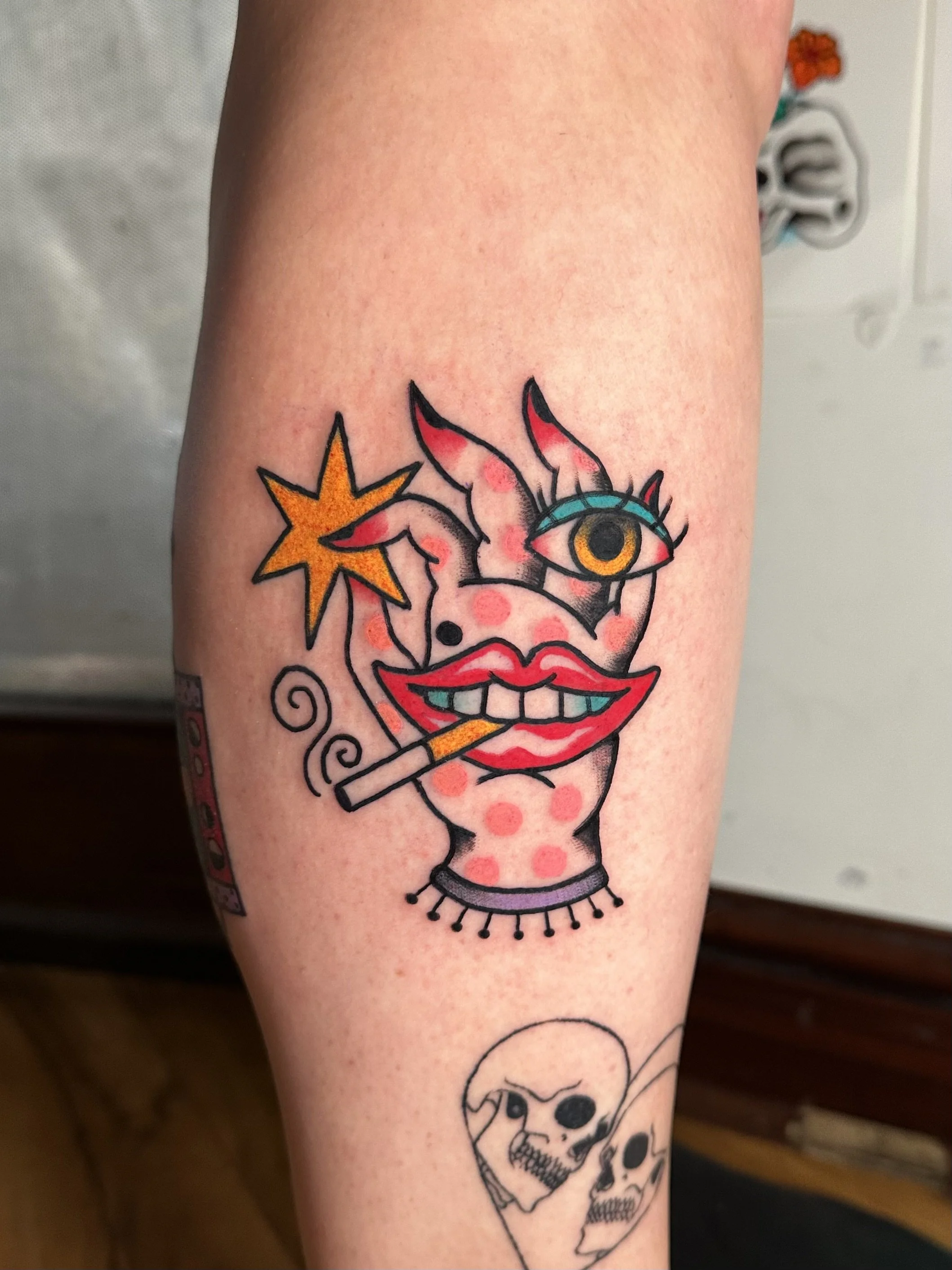 A colorful tattoo featuring a surreal clown face with exaggerated red lips, a cigarette, a star, an eye, and dotted patterns with another smaller tattoo of skulls below.