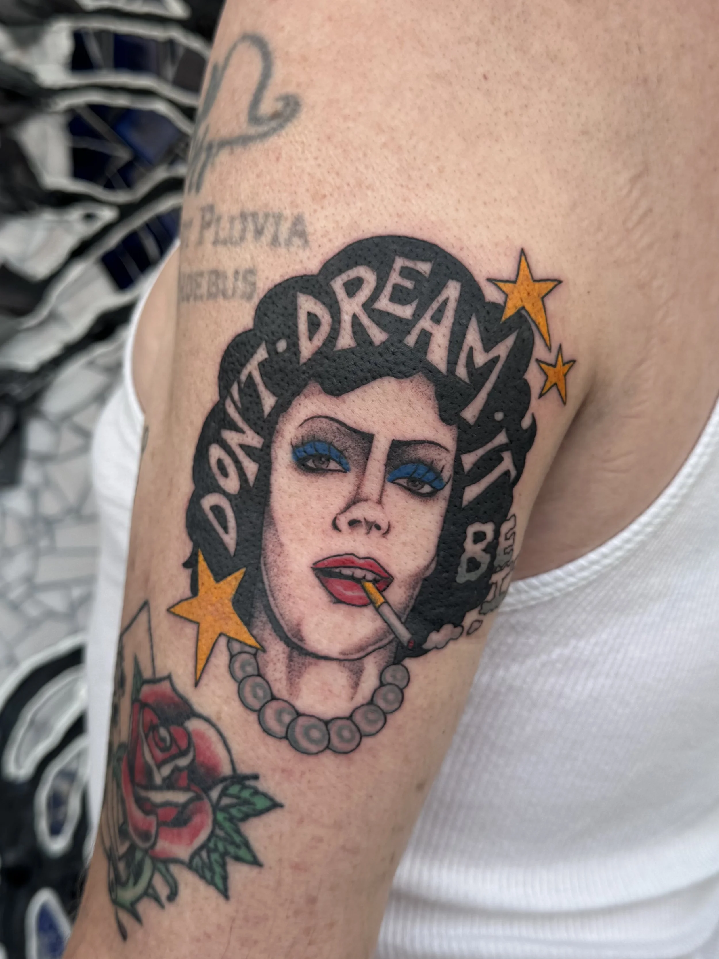 Tattoo of Dr. Frankenfurter from Rocky Horror Picture Show with blue eyeshadow, red lipstick, smoking a cigarette, with the words 'Don't Dream It' in his hair surrounded by yellow stars, and a tattoo of a rose on her arm.