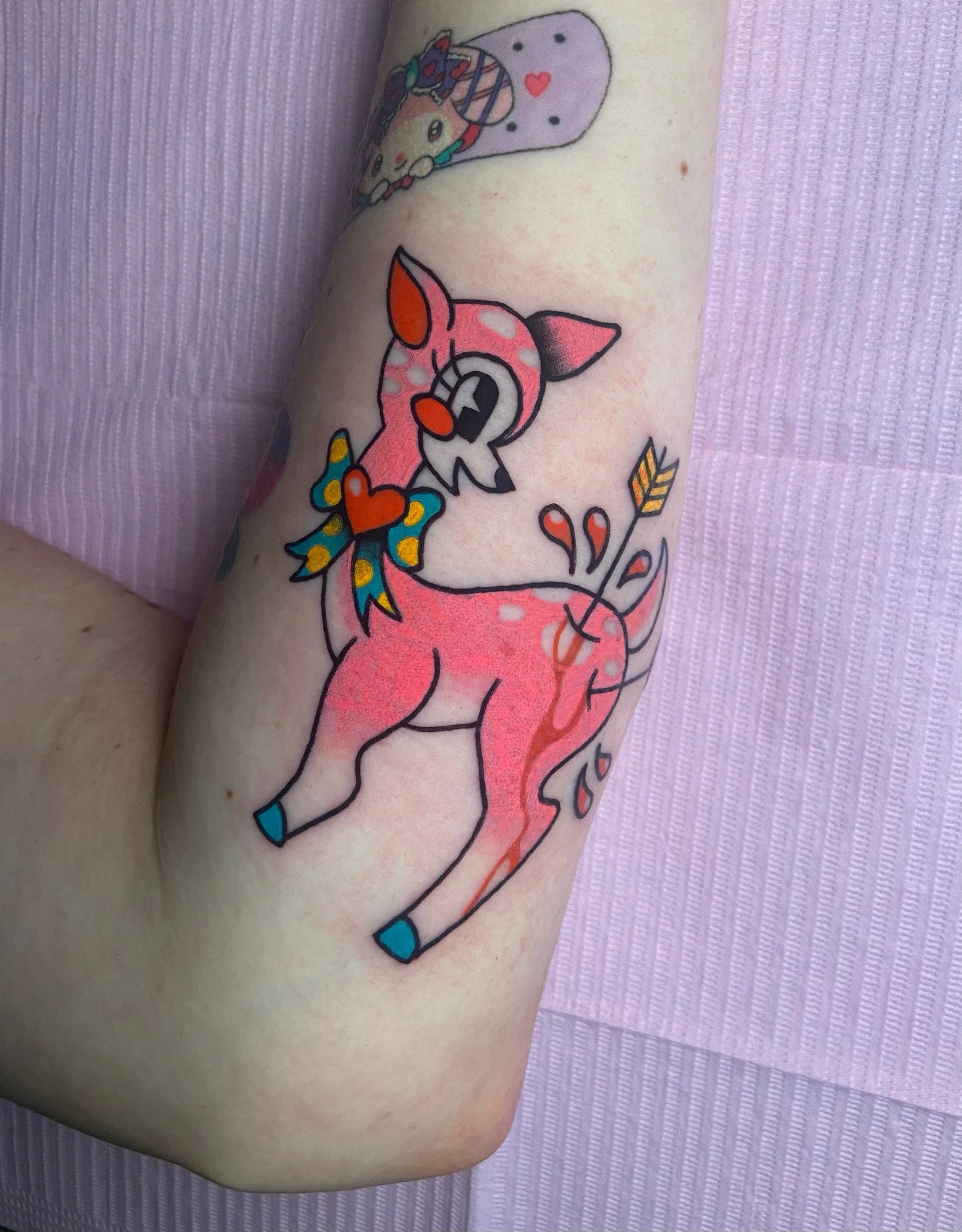 A colorful cartoon-style tattoo of a pink deer with a bow around its neck, hearts, and arrows on its body, on a person's arm.