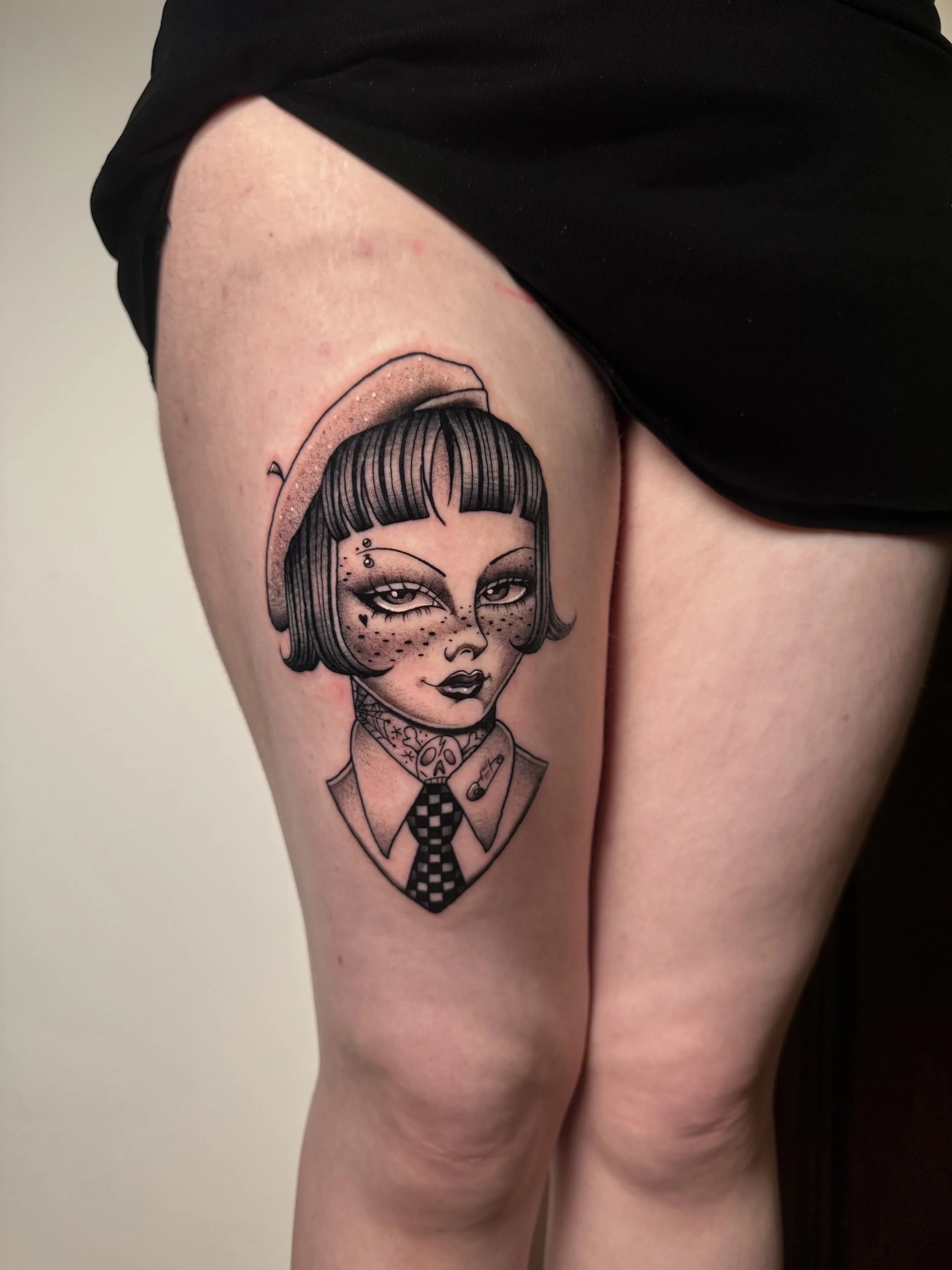Tattoo of a stylized woman with short hair, freckles, and makeup, wearing a hat, collared shirt, and a checked tie, with a tattoo of a needle above her eye on her face.