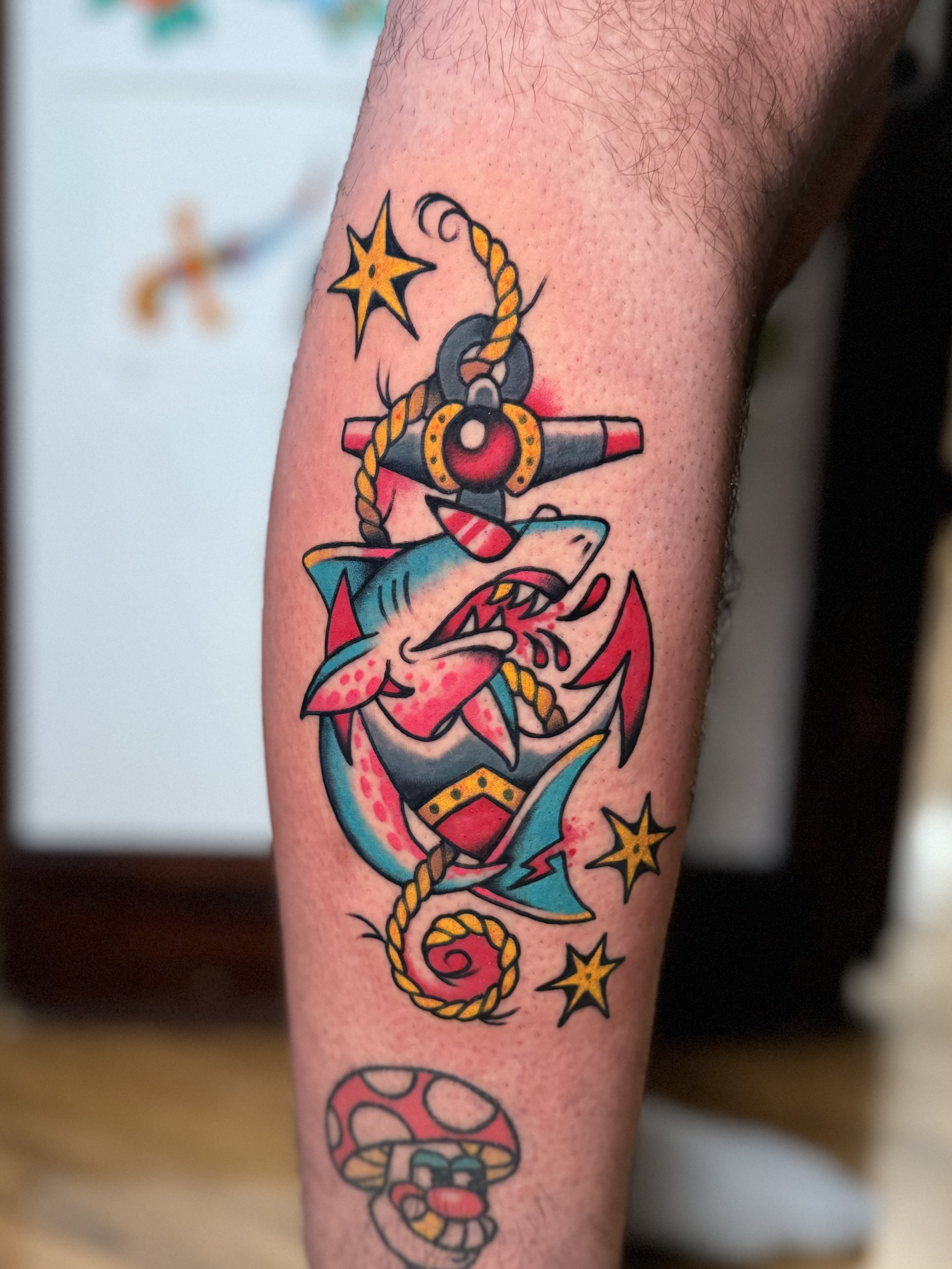 Tattoo of a cartoon-style shark with sunglaasses on with an anchor surrounded by stars, an anchor, and a mushroom at the bottom.
