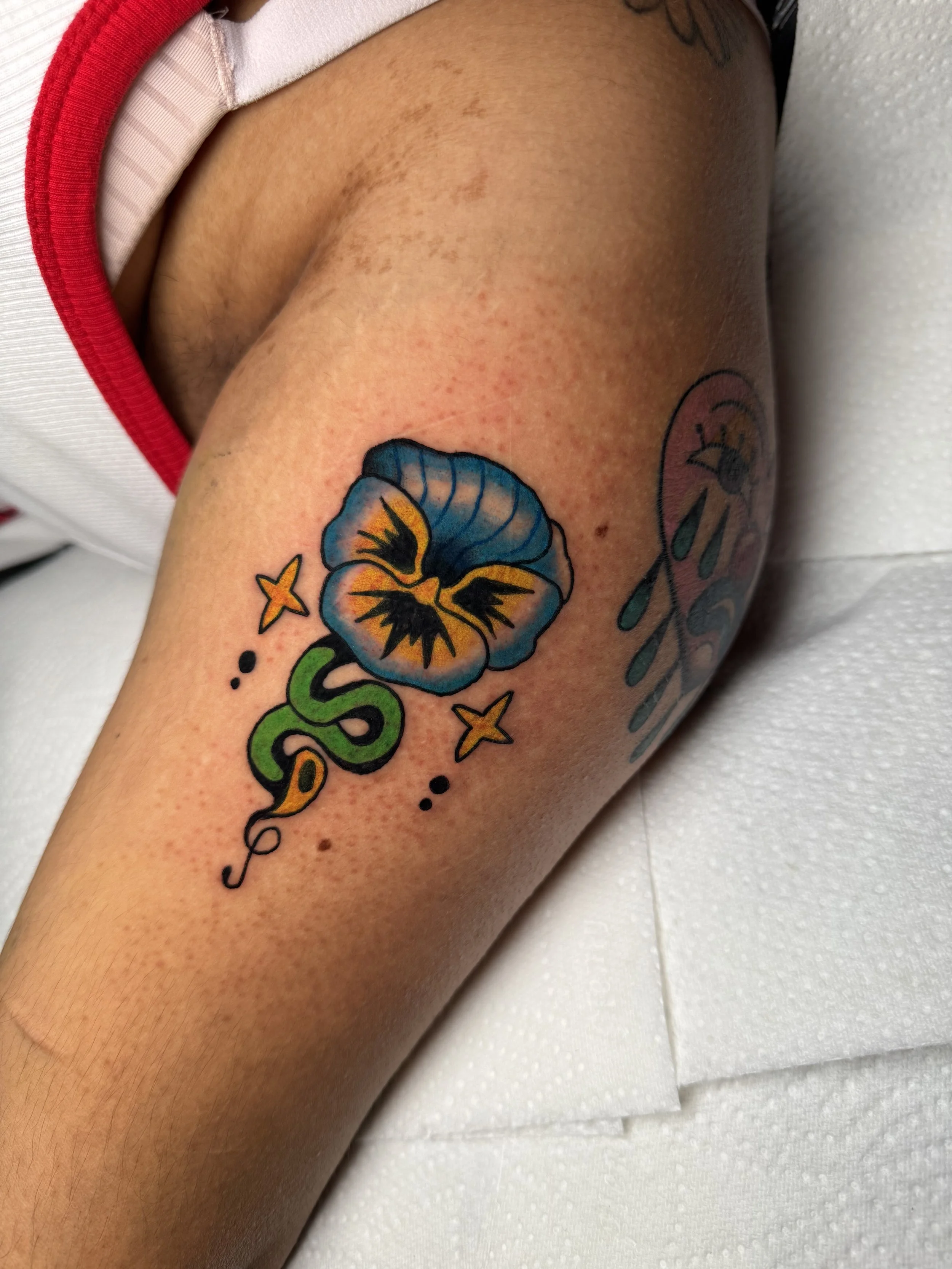 Colorful tattoo of a pansy flower with green lettering and yellow stars on the person's upper arm.