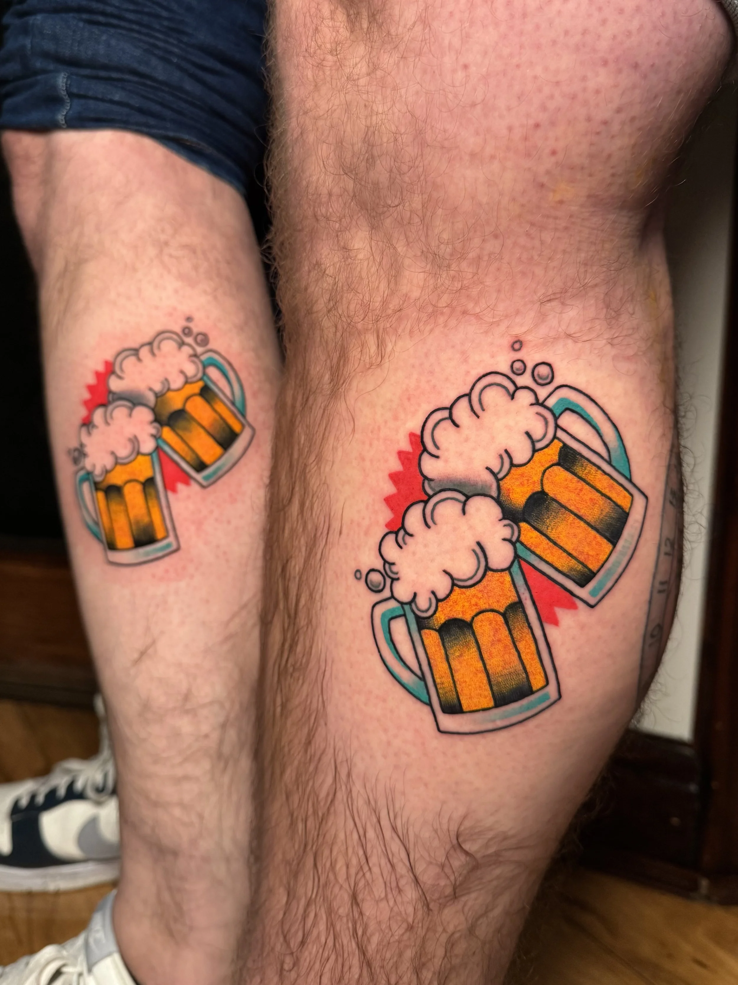Two tattoos of beer mugs clinking together, with foam spilling over and a red splash behind them.