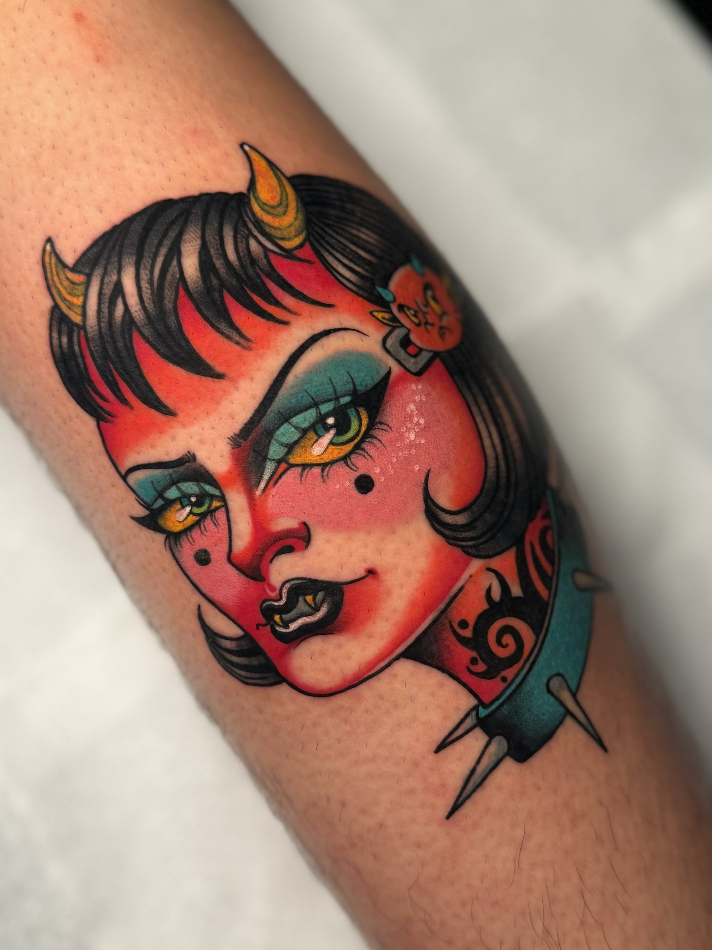 Colorful tattoo of a devil woman's face with punk hairstyle, makeup, small horns, a tattooed neck, and a spiked collar.
