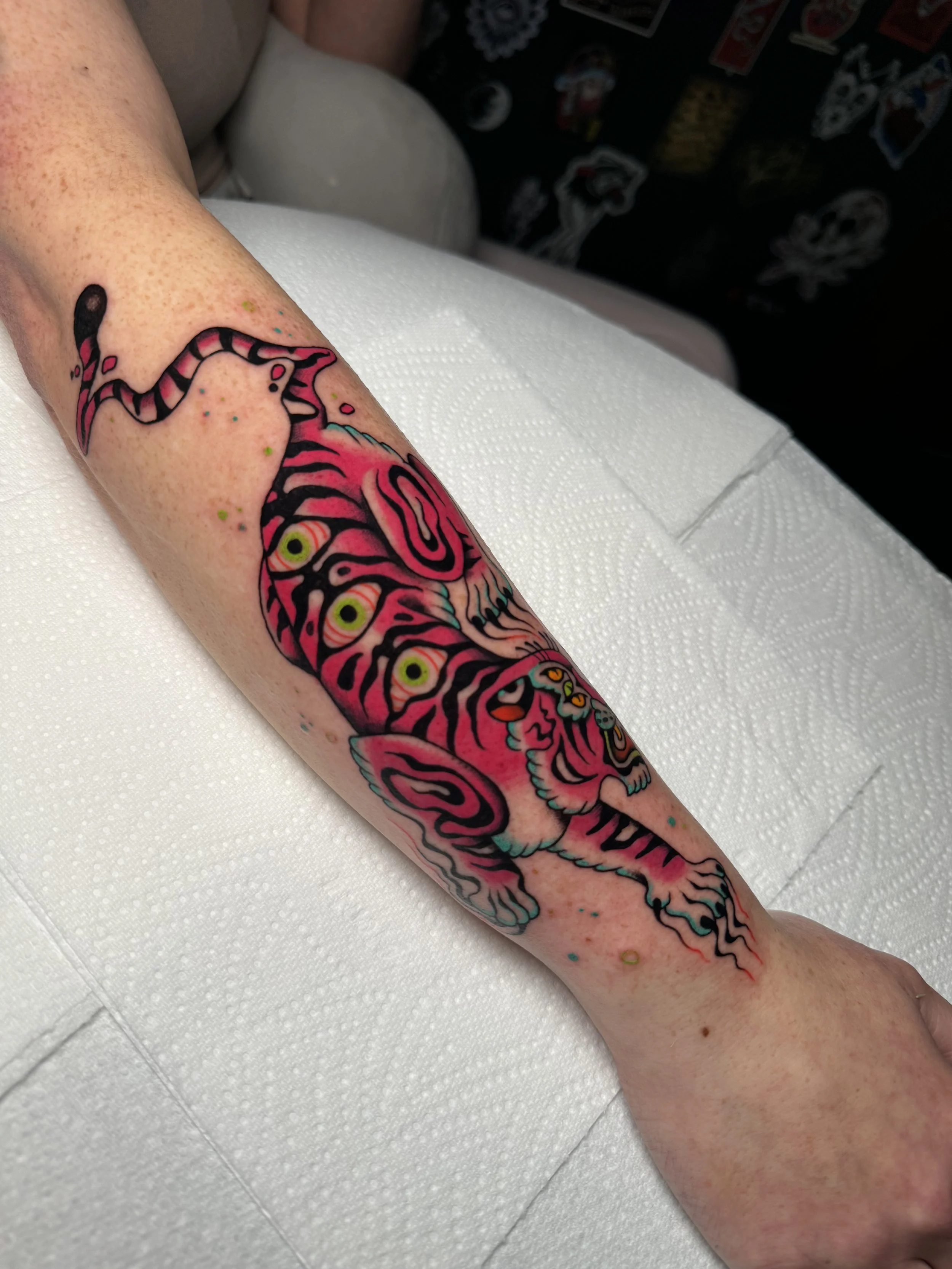 Colorful tattoo of a tiger on a person's forearm, featuring pink, black, yellow, and teal colors, with a stylized design and multiple eyes on the tiger's face.