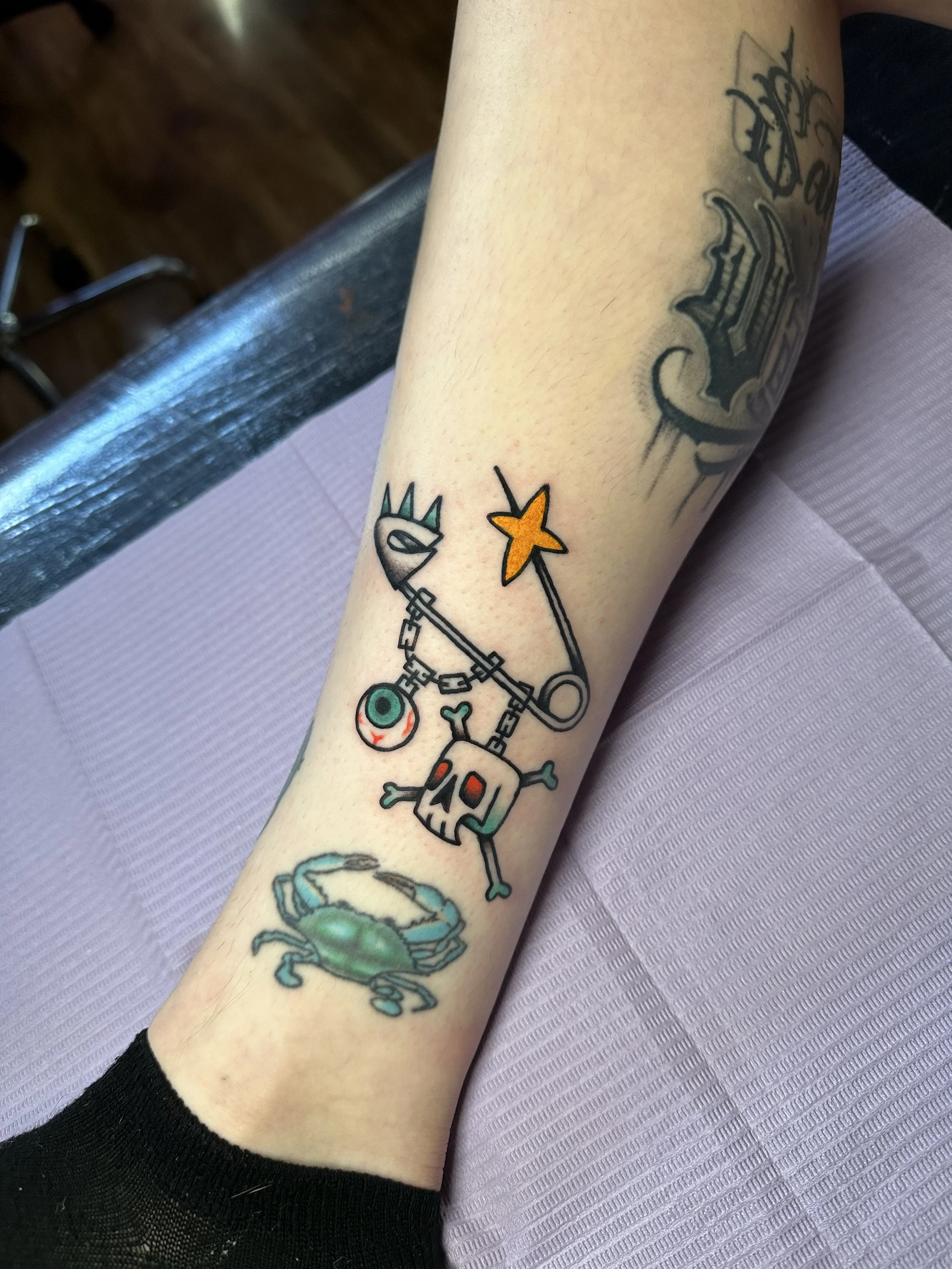 Tattoo of a cartoonish skull with crossbones, a star, and a chain, attached to a punk style safety pin on a person's ankle.