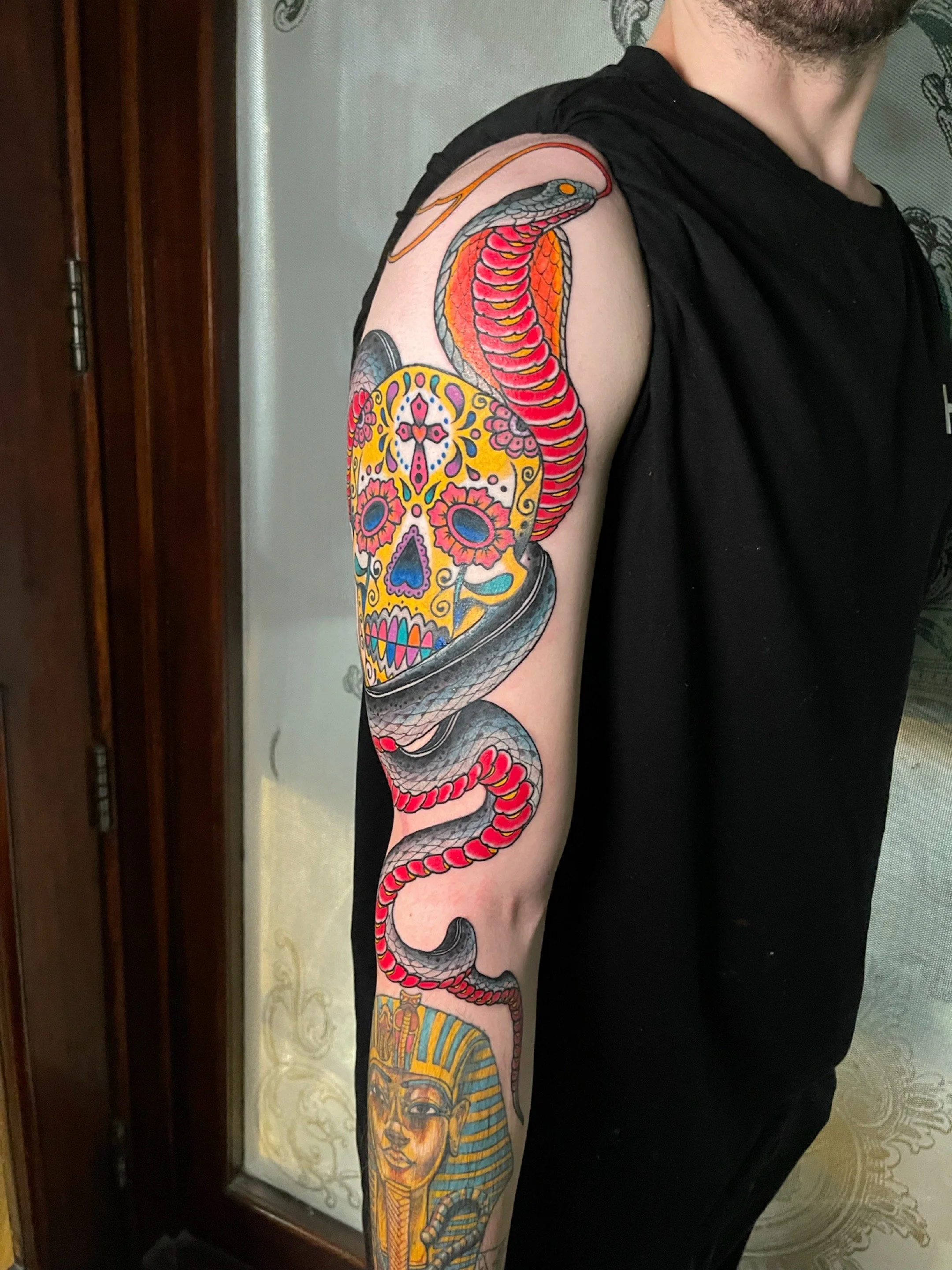 Tattoo of a colorful sugar skull with floral designs on a person's upper arm, intertwined with a detailed cobra snake with red and black markings.