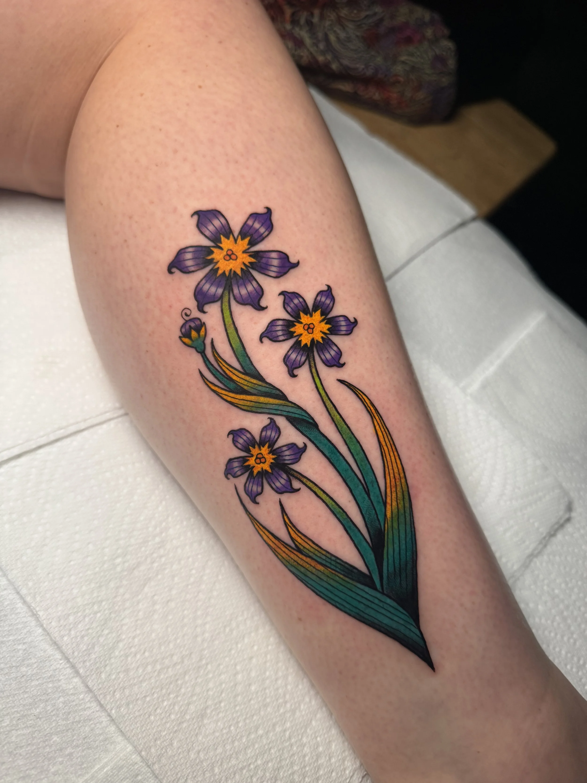 Colorful tattoo of purple flowers (bermudianas) with yellow centers and green leaves on person's lower leg.