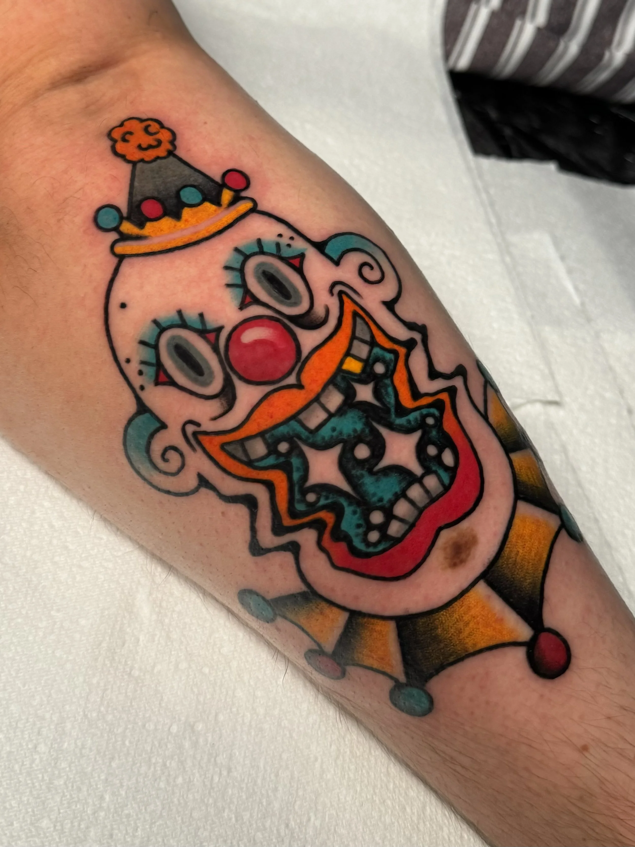 Colorful traditional tattoo of a clown face with a jester hat, exaggerated facial features, and vibrant colours.