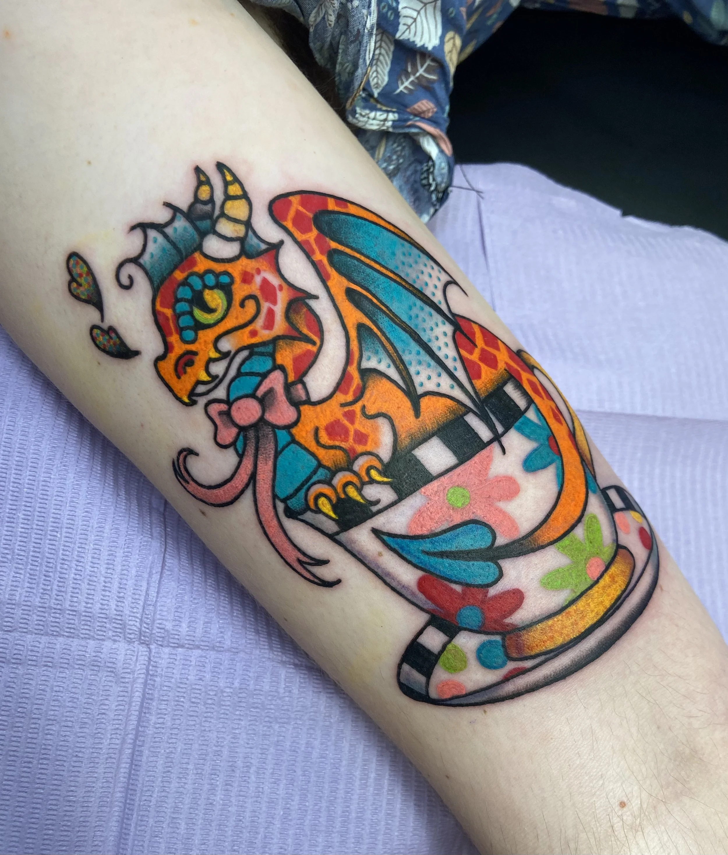 Colorful tattoo of a dragon with wings inside a tea cup on a person's arm.