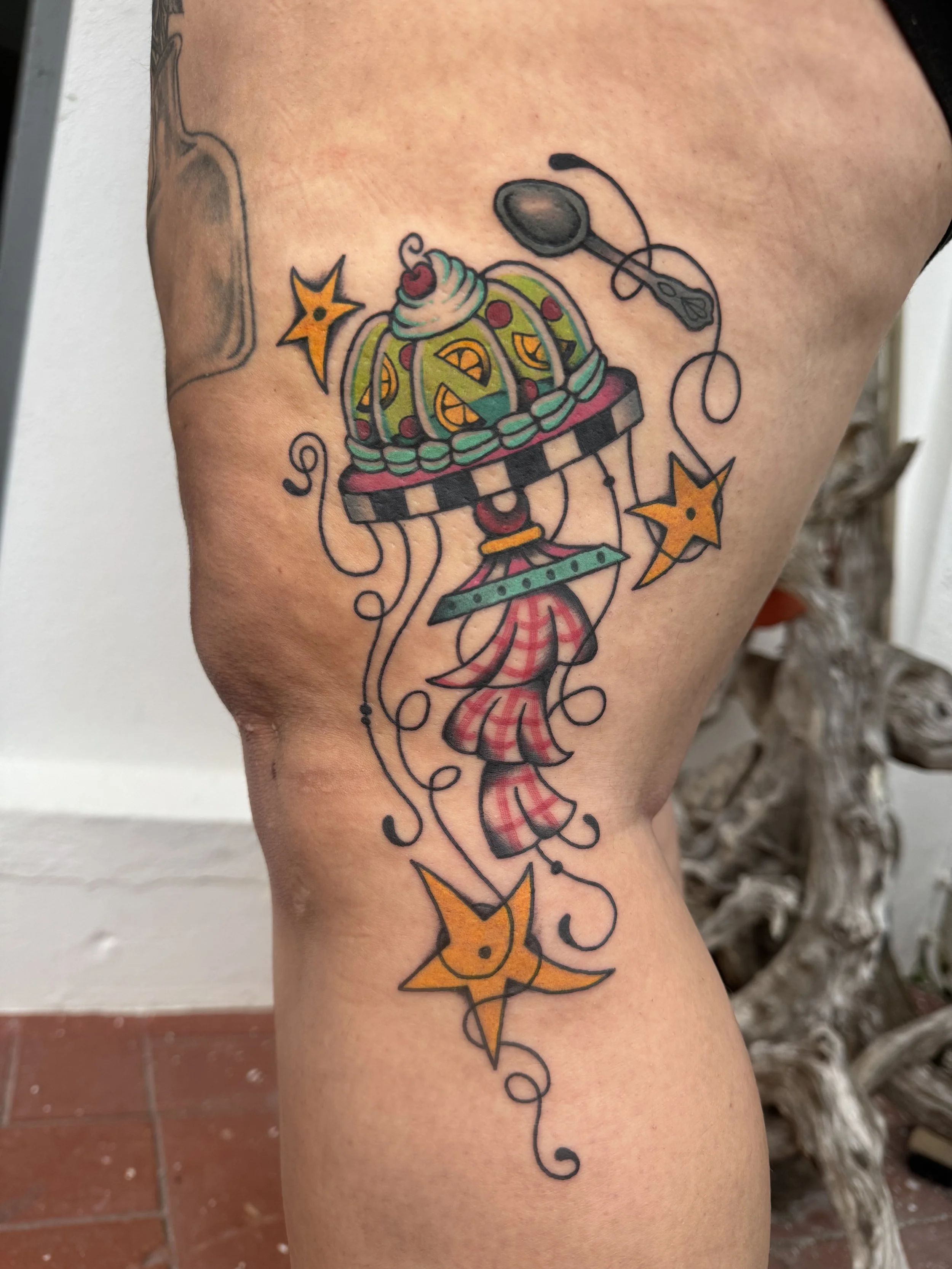 Colorful tattoo of a crown, stars, a cupcake, and a hanging dress on upper thigh.