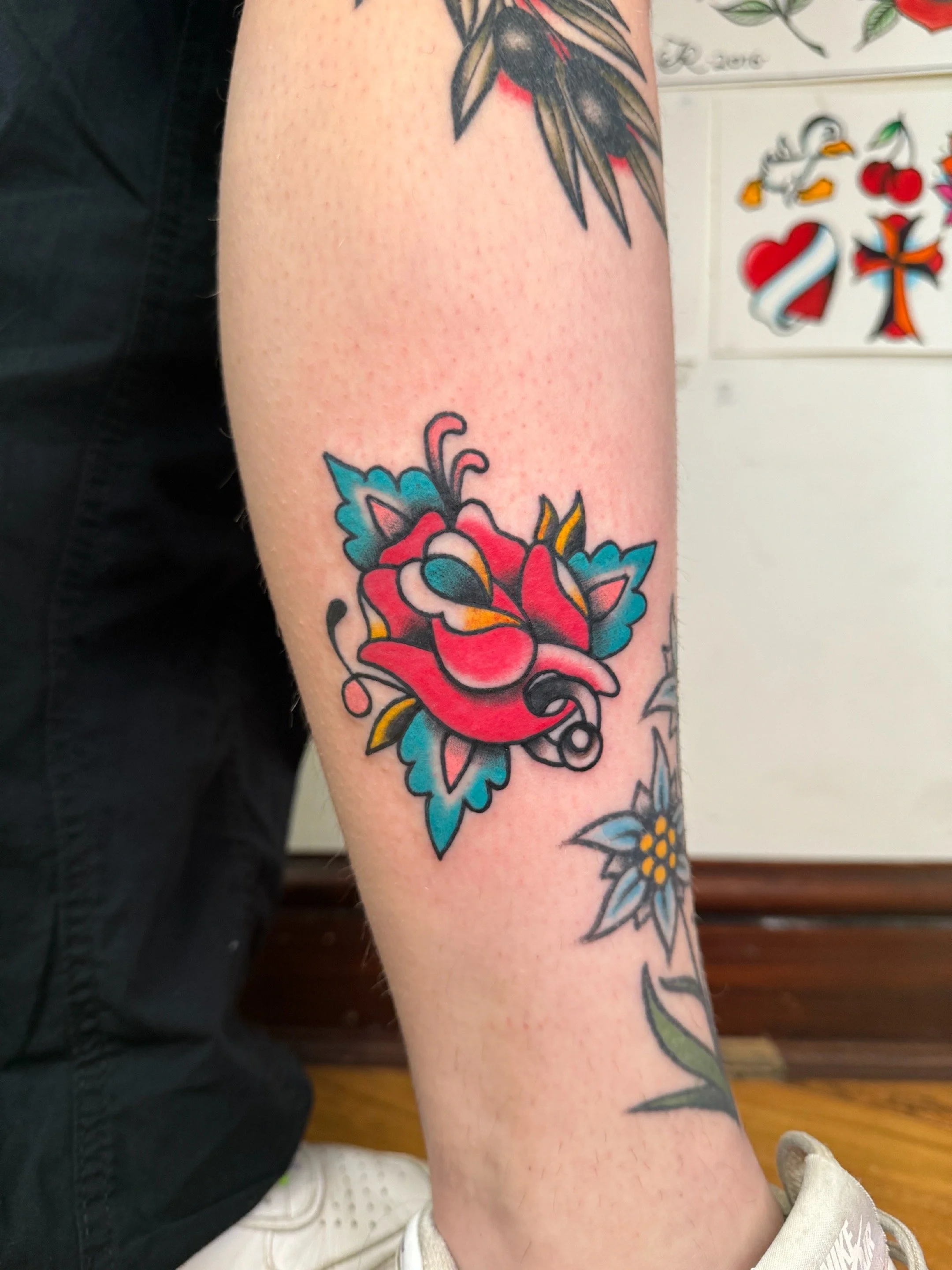 Colorful tattoo of a red rose with green and blue leaves on a person's forearm.