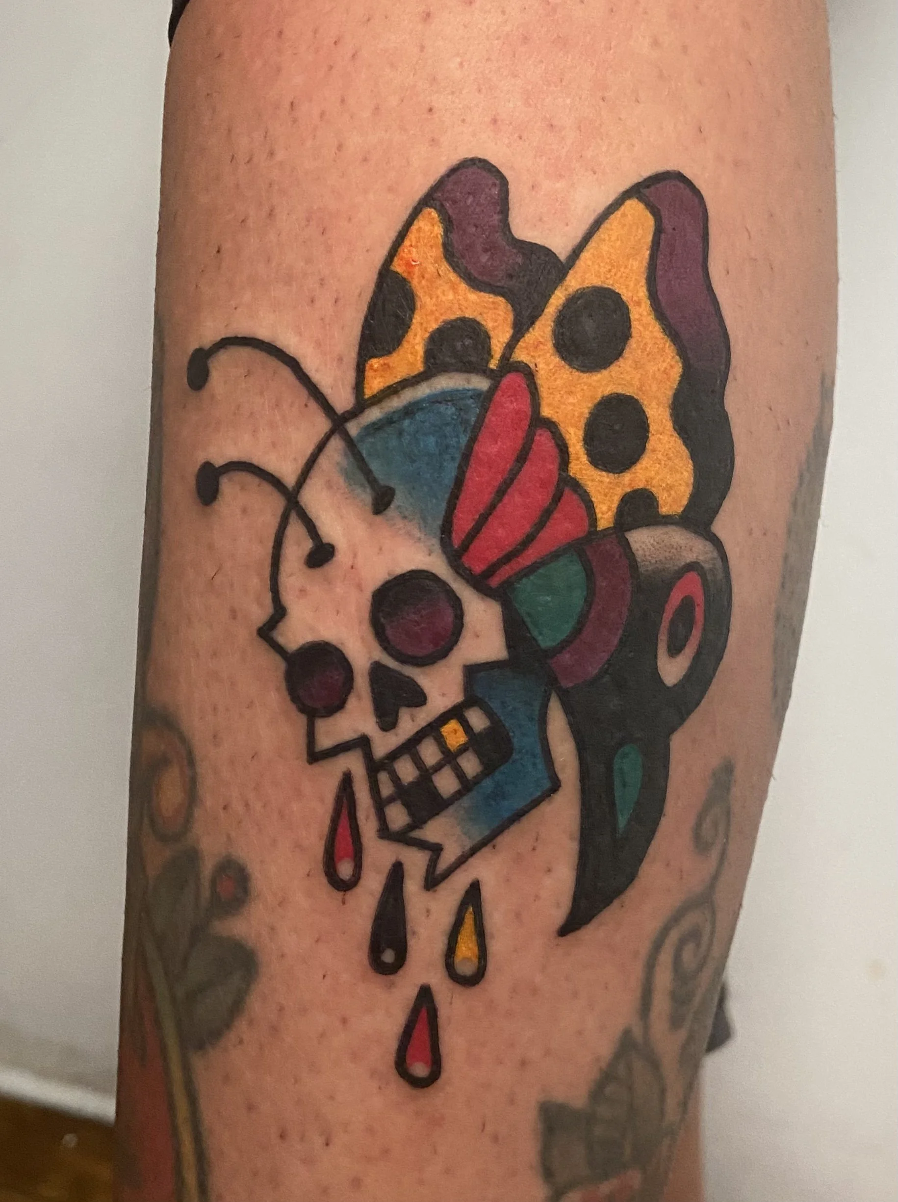 Colorful tattoo of a skull with butterfly wings, a tear, and dripping blood.
