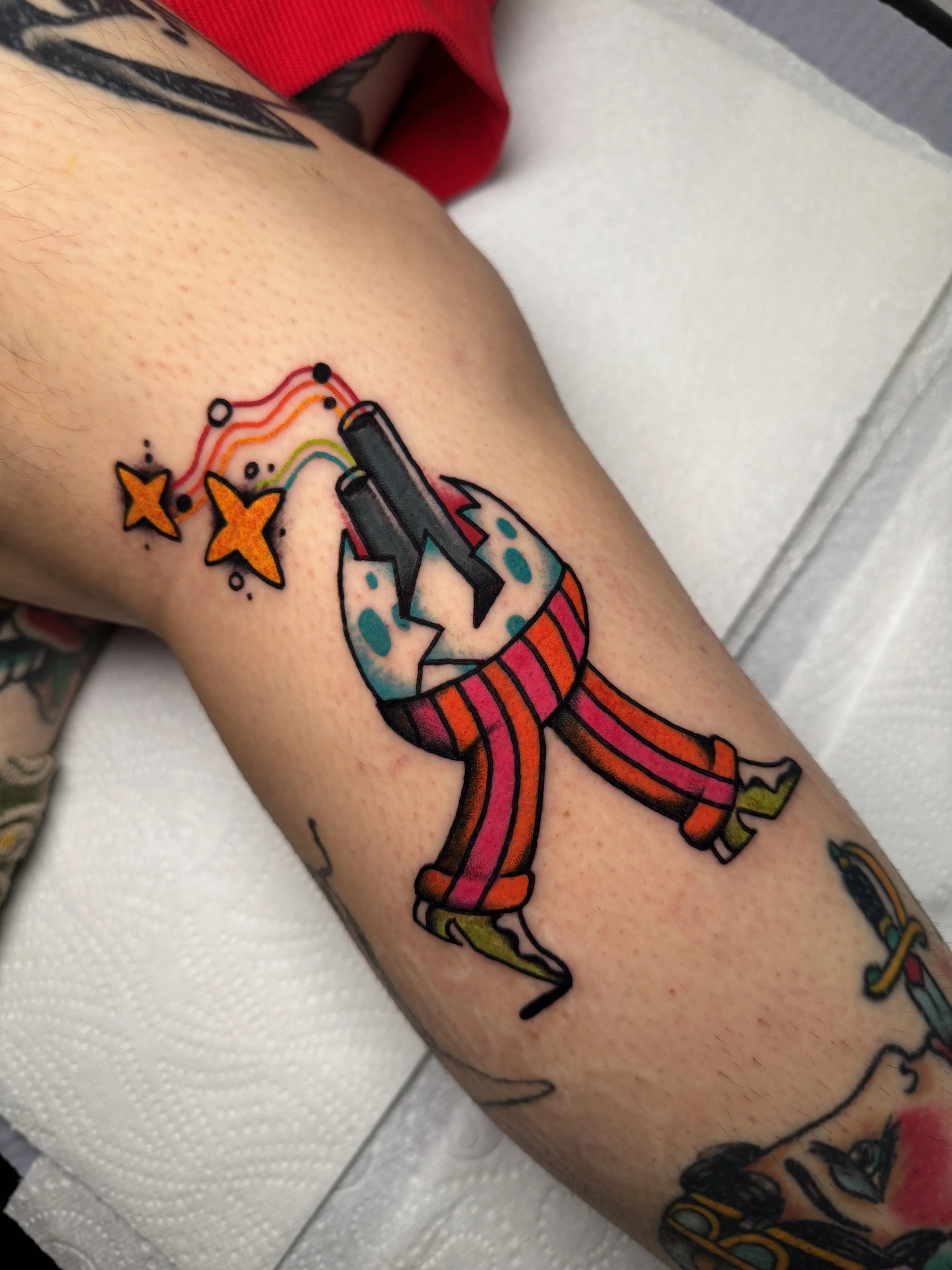 Colorful tattoo of a psychedelic egg in fancy trousers and green shoes with a smokestack peaking out of it, pumping rainbows and sparkles into the air.