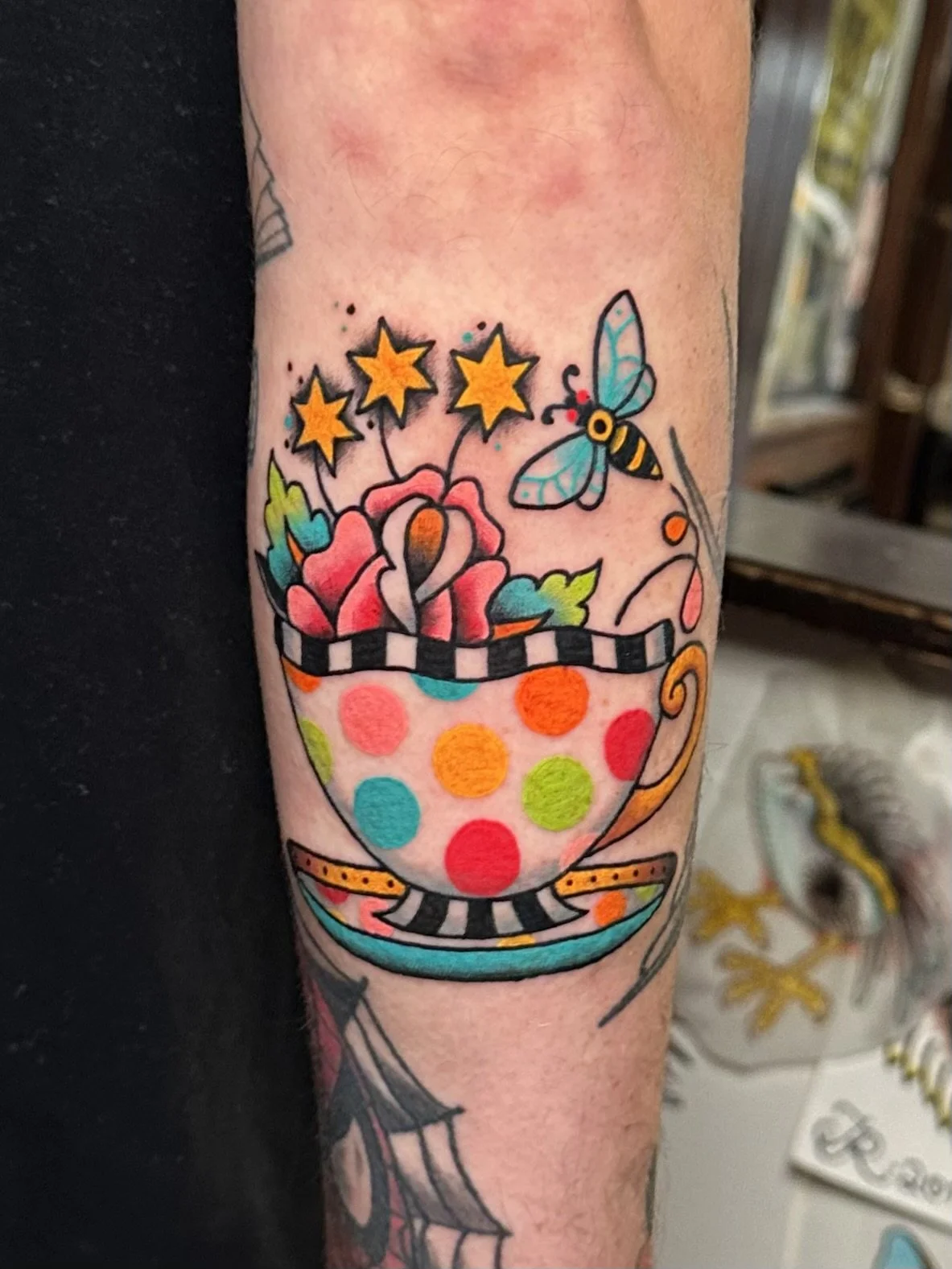 Colorful tattoo of a teacup filled with flowers, a dragonfly, and stars, featuring polka dots, stripes, and vibrant colors.
