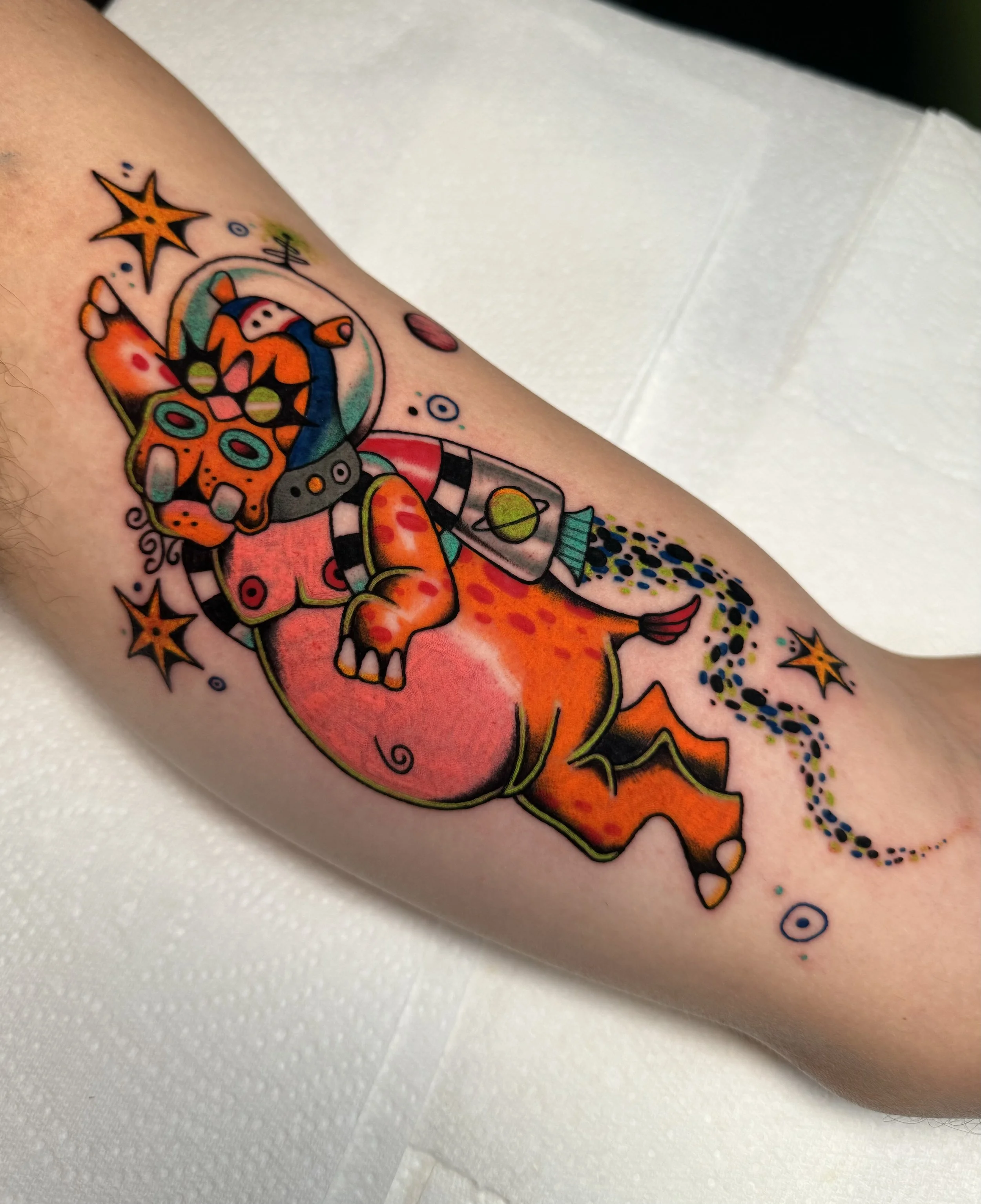 Colorful tattoo of a cartoon hippo dressed as an astronaut with a jetpack with stars and planets around it on a person's arm.