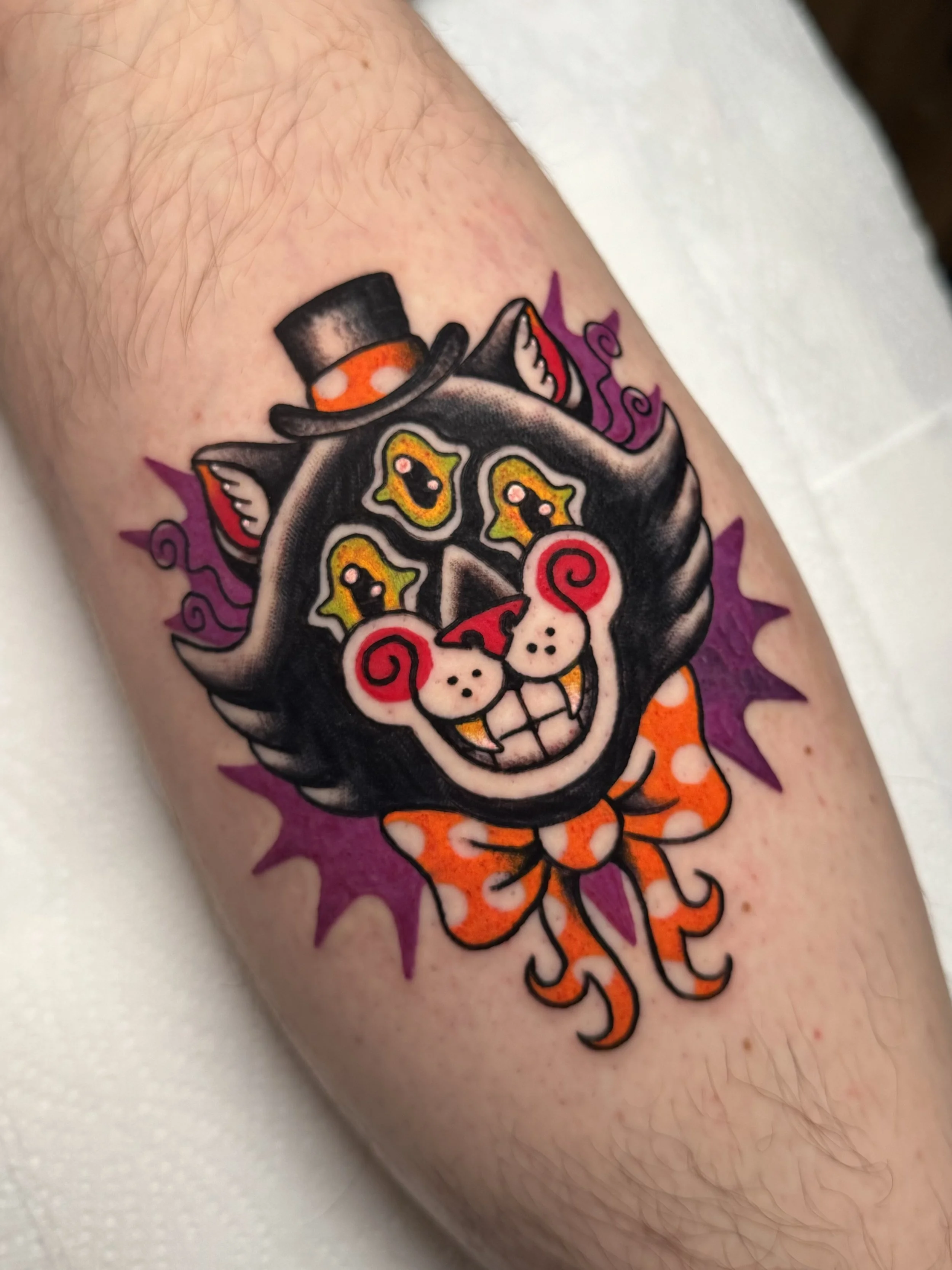 Colorful tattoo of a grinning black cat with yellow eyes, red spiraled cheeks, wearing an orange bow with white polka dots, a black top hat with an orange band, and a purple splash background.